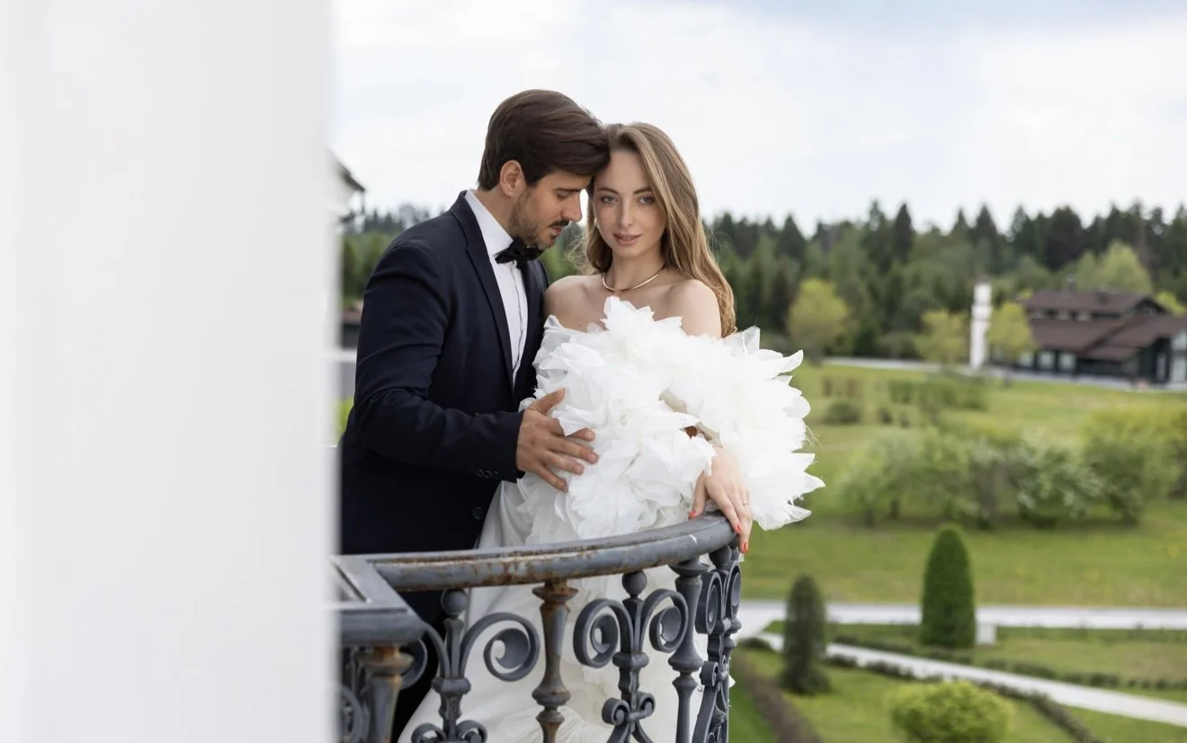 A man in a black tuxedo and a woman in a white wedding dress stand on a balcony, holding a large bouquet of white flowers and looking at each other, with a lush green landscape in the background.