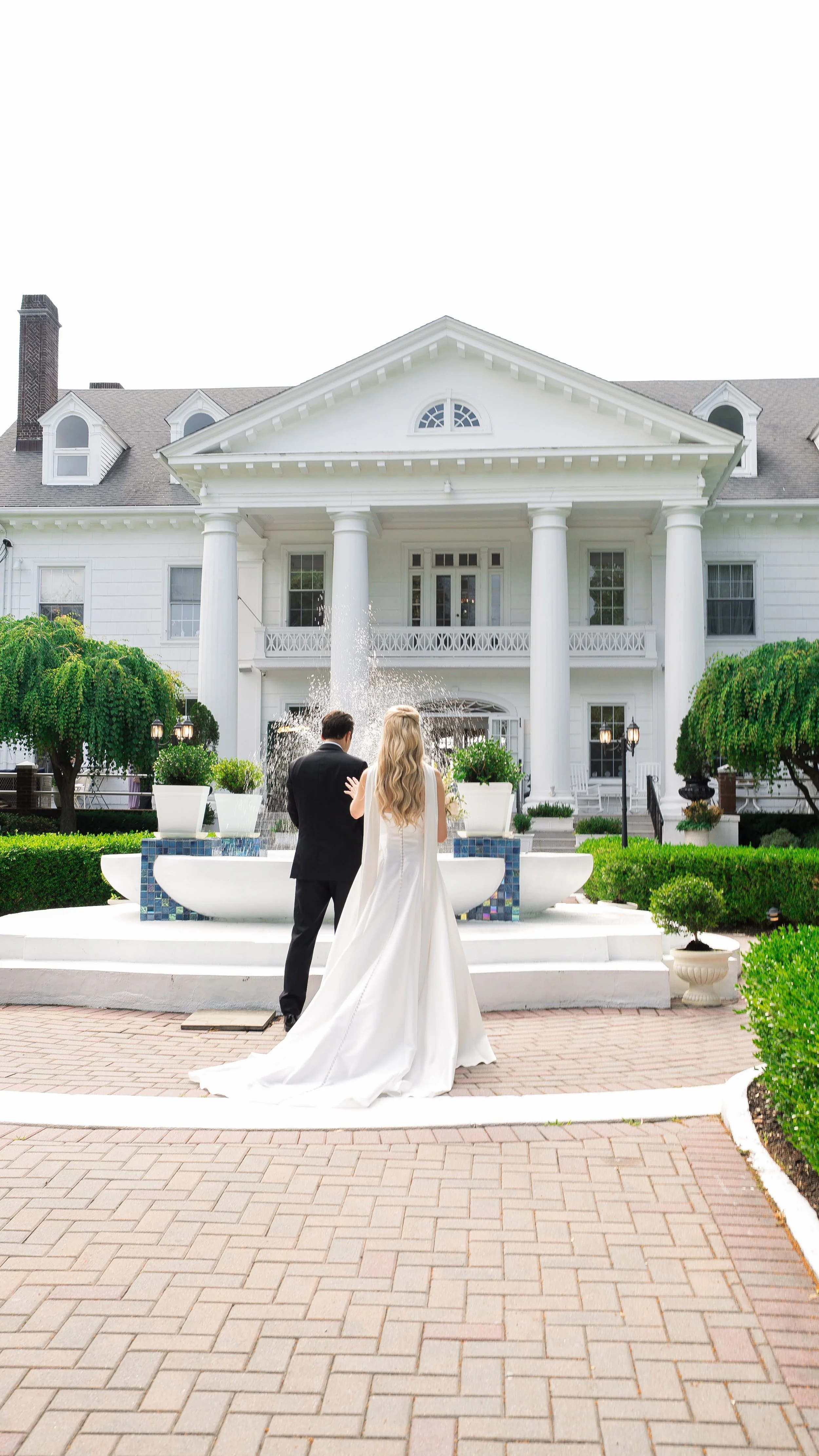 A bride and groom stand together in front of a large white house with four prominent columns, a pent roof, and a fountain. The bride wears a long white wedding dress, and the groom is dressed in a black suit. Green trees and manicured bushes adorn th
