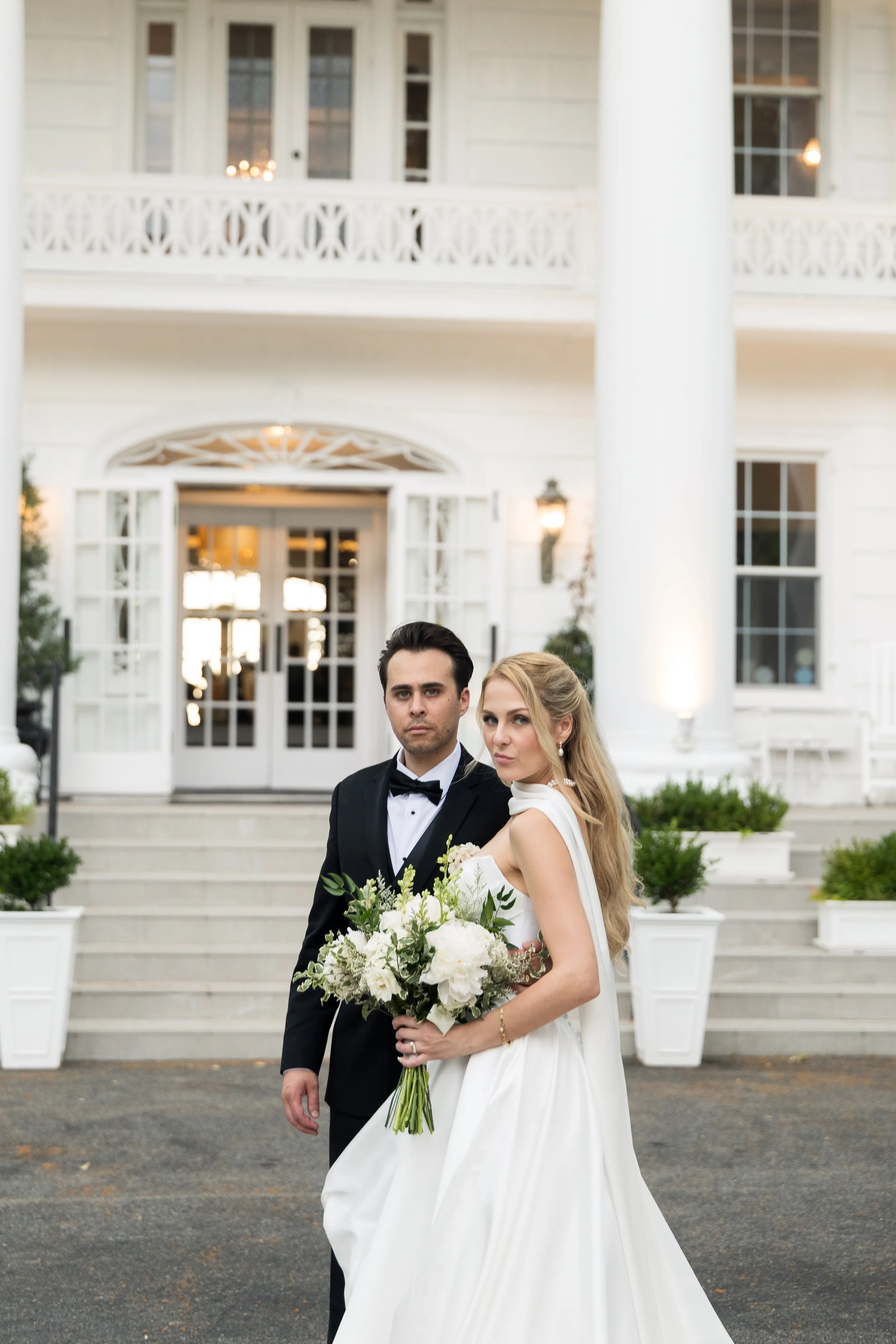 A newlywed couple stands in front of a large white house with a staircase and potted plants. The groom is in a tuxedo, and the bride is in a white wedding gown holding a bouquet of white flowers with greenery.