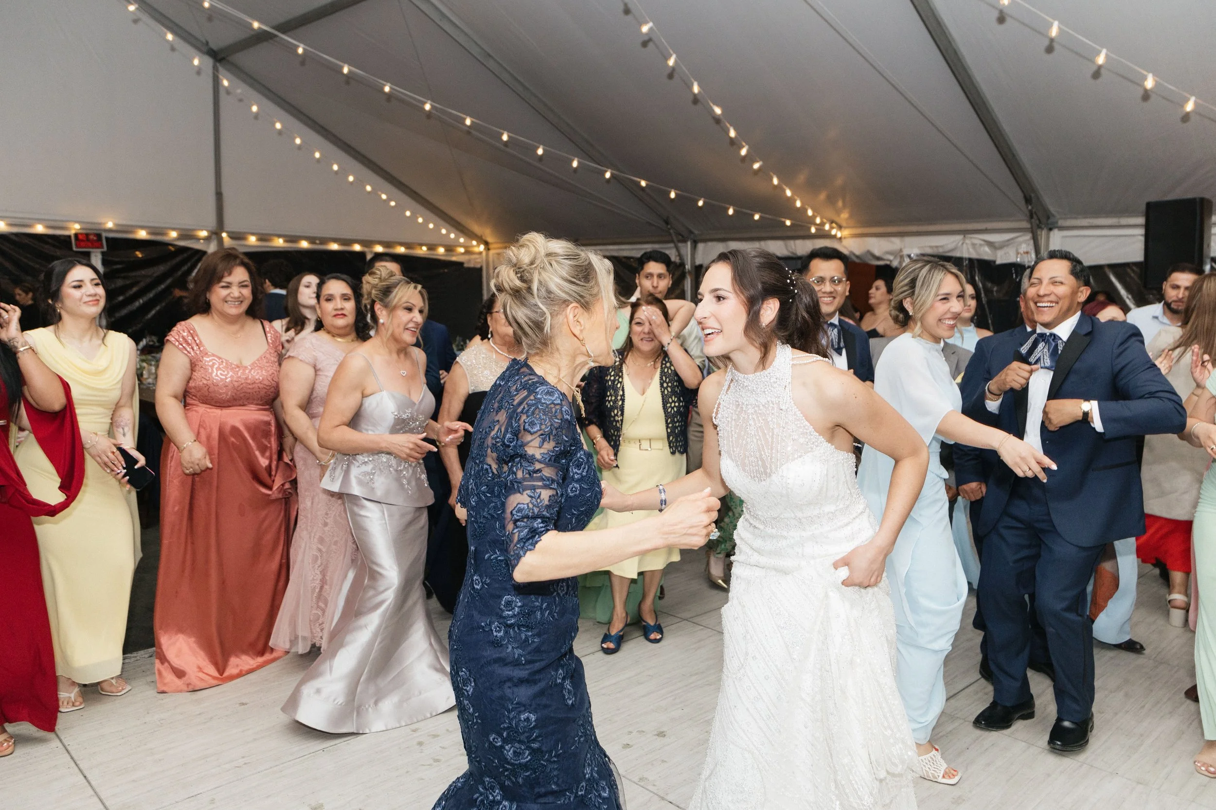 People dancing and celebrating at a wedding reception inside a large tent decorated with string lights.