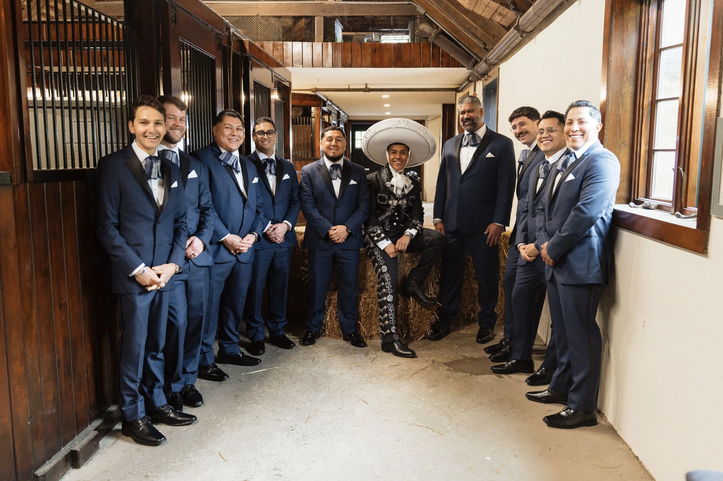 Group of eleven men and women dressed in formal suits, standing in a barn-like setting with wooden walls and hay bales, smiling for the photo.