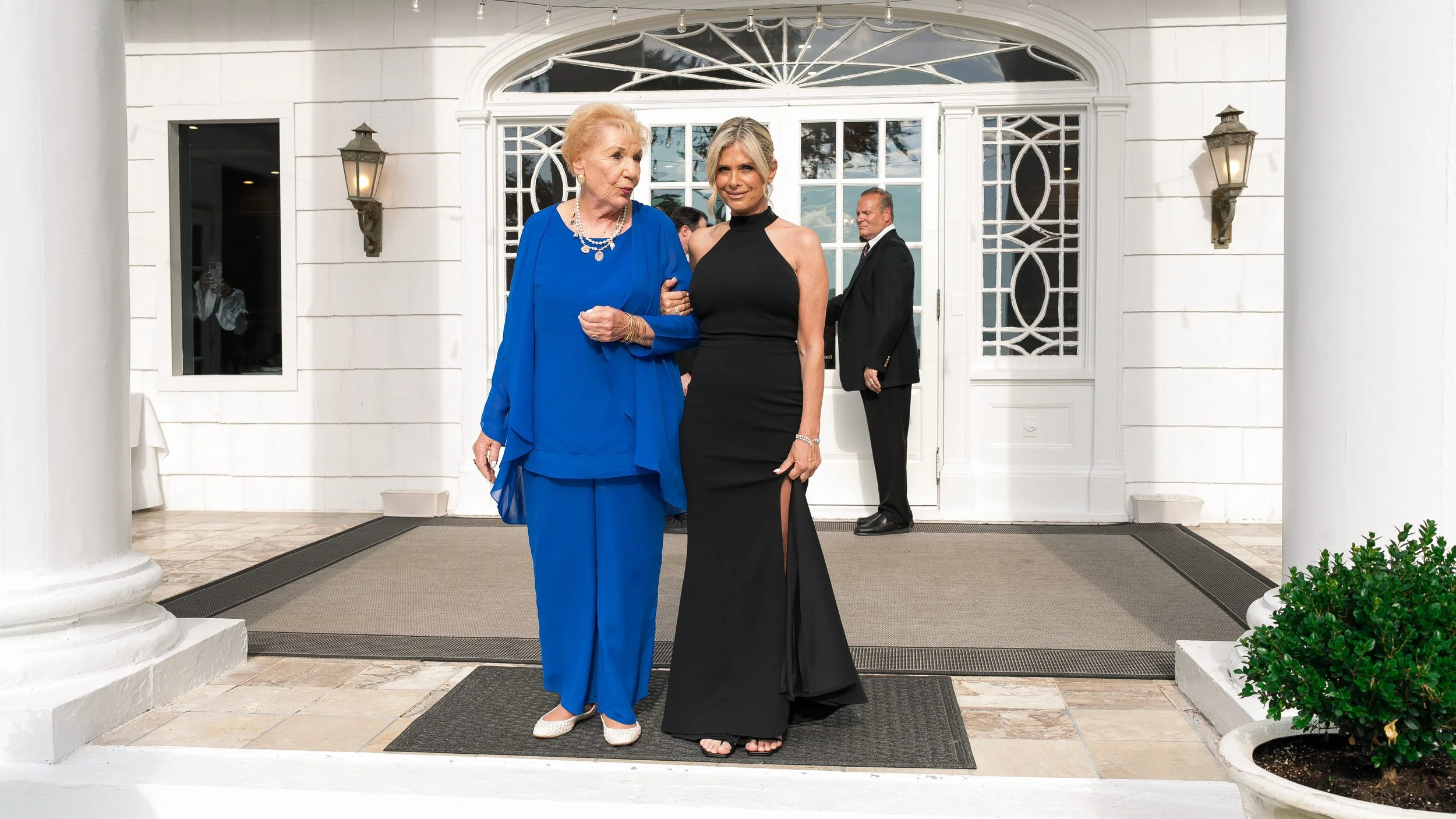 Two women standing arm in arm outside a white building with a glass door, one in a bright blue dress with jewelry and the other in a black evening gown, with a man in a suit in the background.