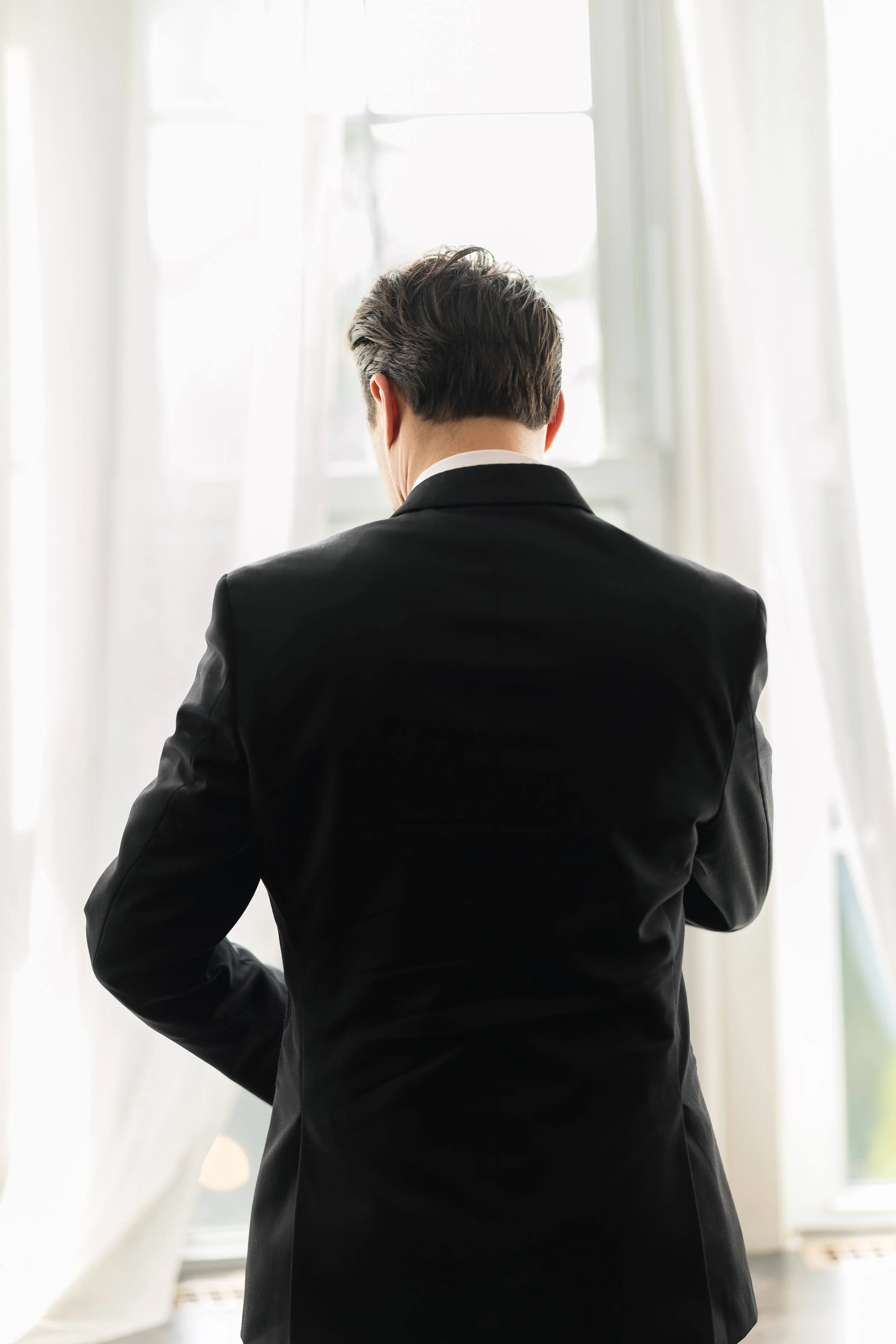 Back view of a man in a black suit standing indoors near a window with white curtains, looking outside.