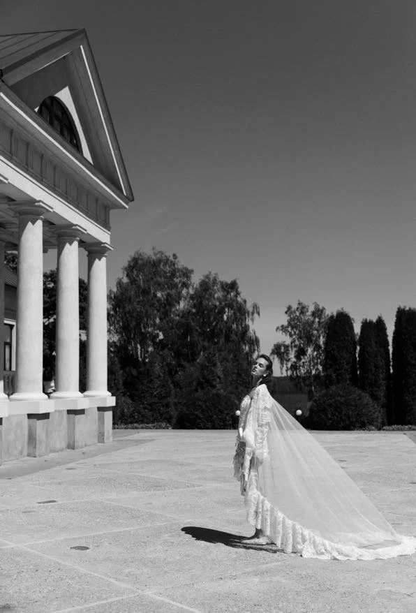 A woman in a wedding dress with a long train stands on an empty outdoor space near a classical building with columns, with trees in the background.