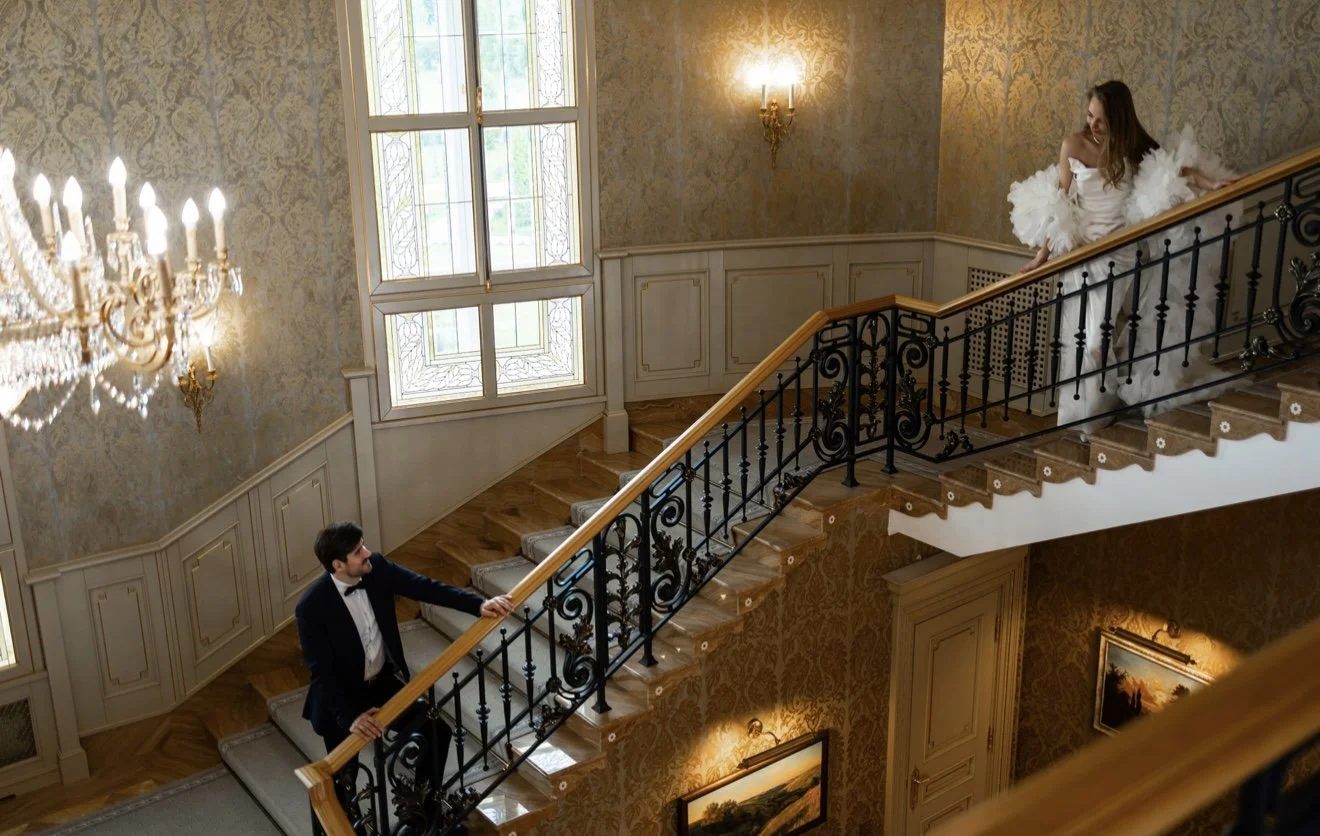 A woman in a white dress and a man in a tuxedo are on a grand staircase in an elegant, chandelier-lit interior. The woman is on the upper level, leaning on the railing, while the man is lower, reaching up toward her.