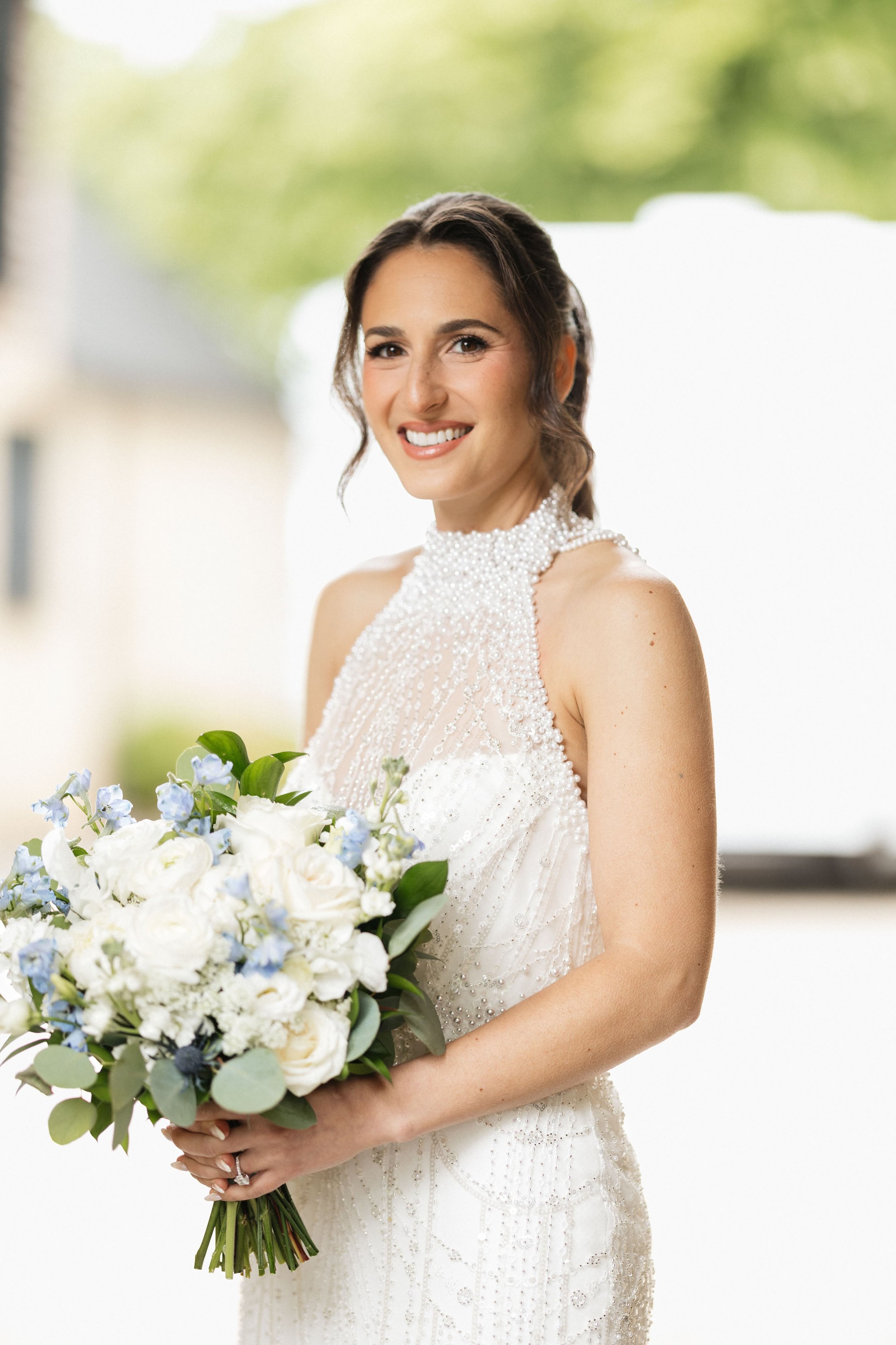 A woman in a white wedding dress holding a bouquet of white and blue flowers, smiling and winking outdoors.