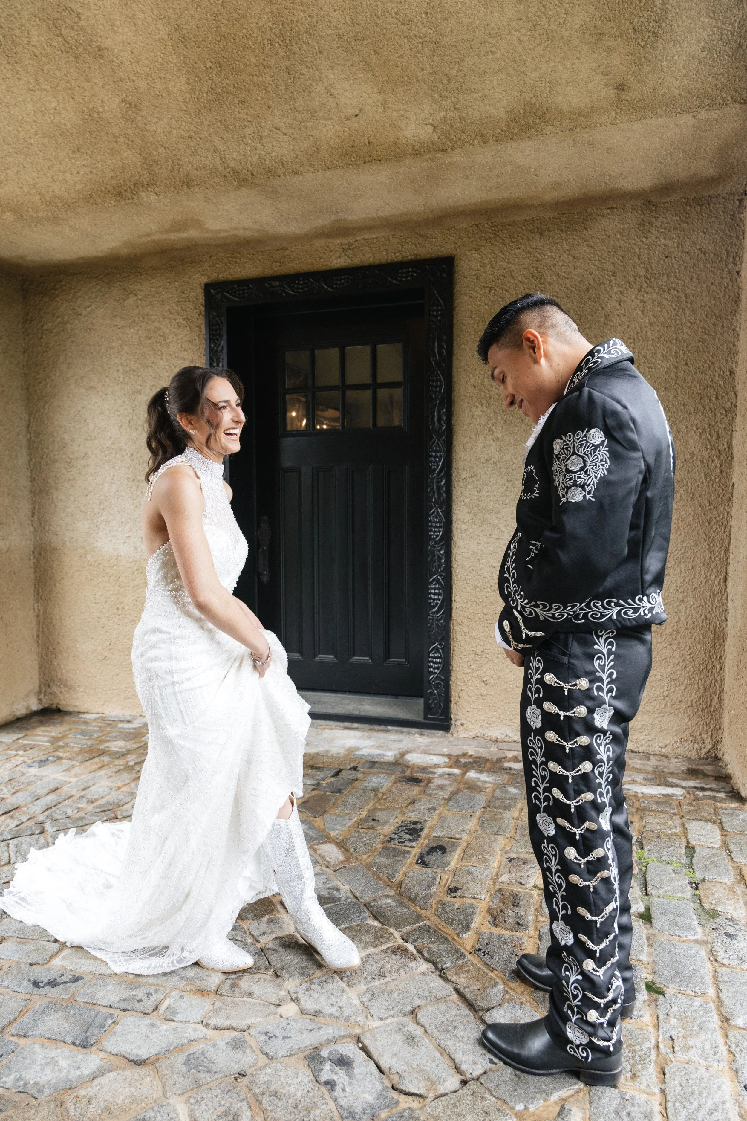 A bride and groom are at their wedding outside on cobblestone pavement. The bride is wearing a white lace wedding dress and white shoes, laughing and holding her dress. The groom is wearing a black suit with white floral embroidery, looking down and 