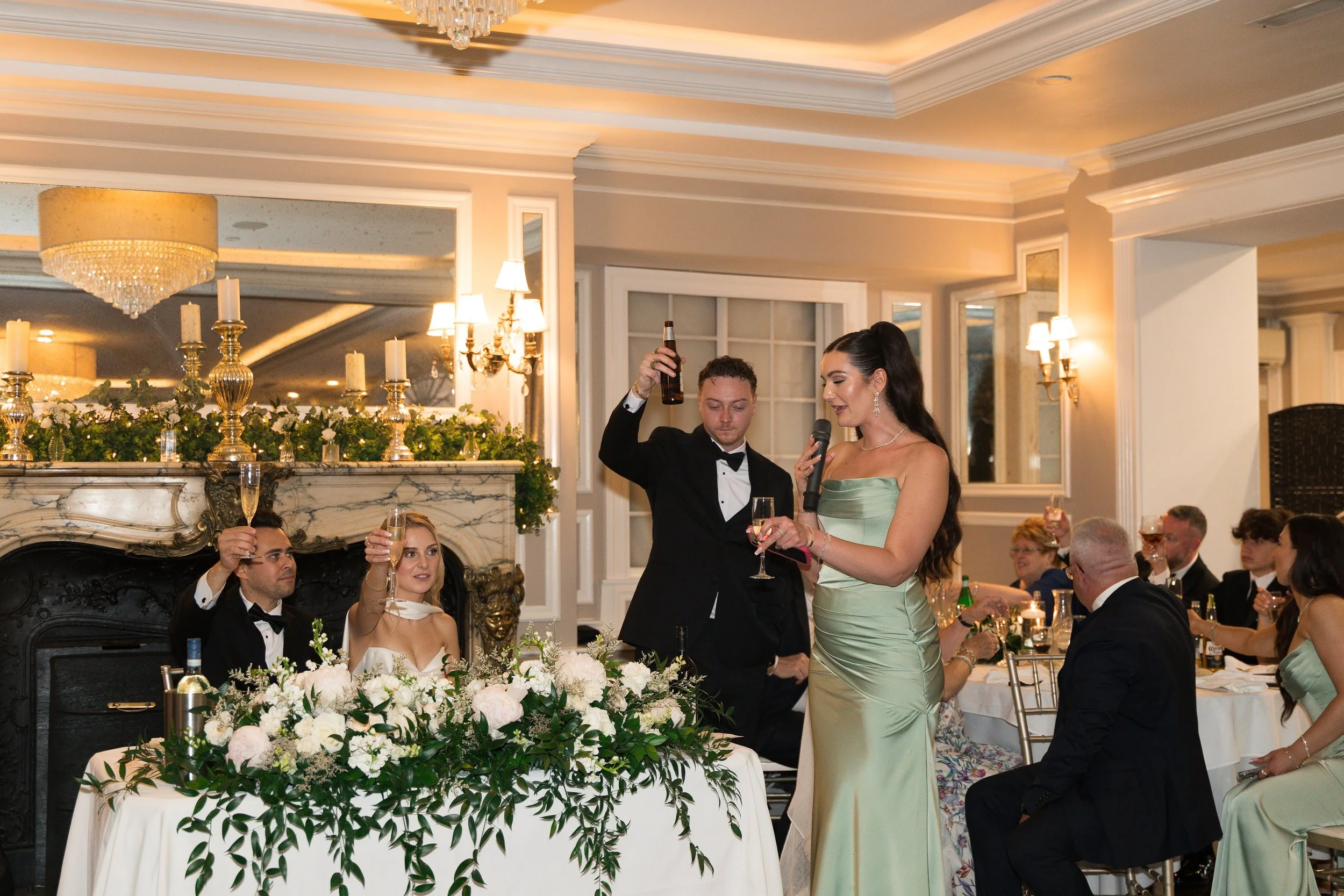 A wedding reception with a bride and groom seated at a head table decorated with white flowers and green foliage. Guests are raising glasses in a toast. The reception is elegant with chandeliers, candles, and warm lighting in a luxurious room.