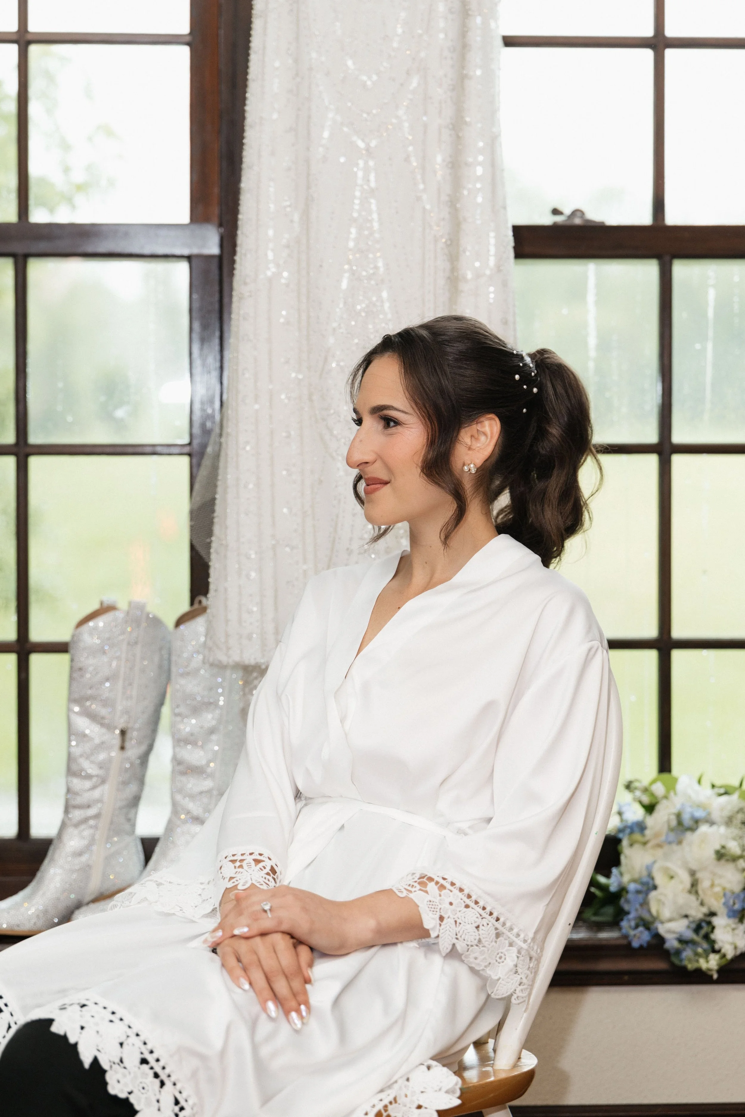 A woman sitting in a white robe, with hair styled in loose waves, seated in front of a window with dark wooden framing. Behind her, wedding shoes and white flower arrangements are visible. The room appears to be a bridal suite or getting ready area.