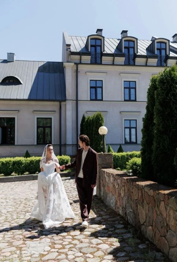 A bride and groom holding hands while walking outside in front of a large, elegant building with a stone wall and lush greenery.