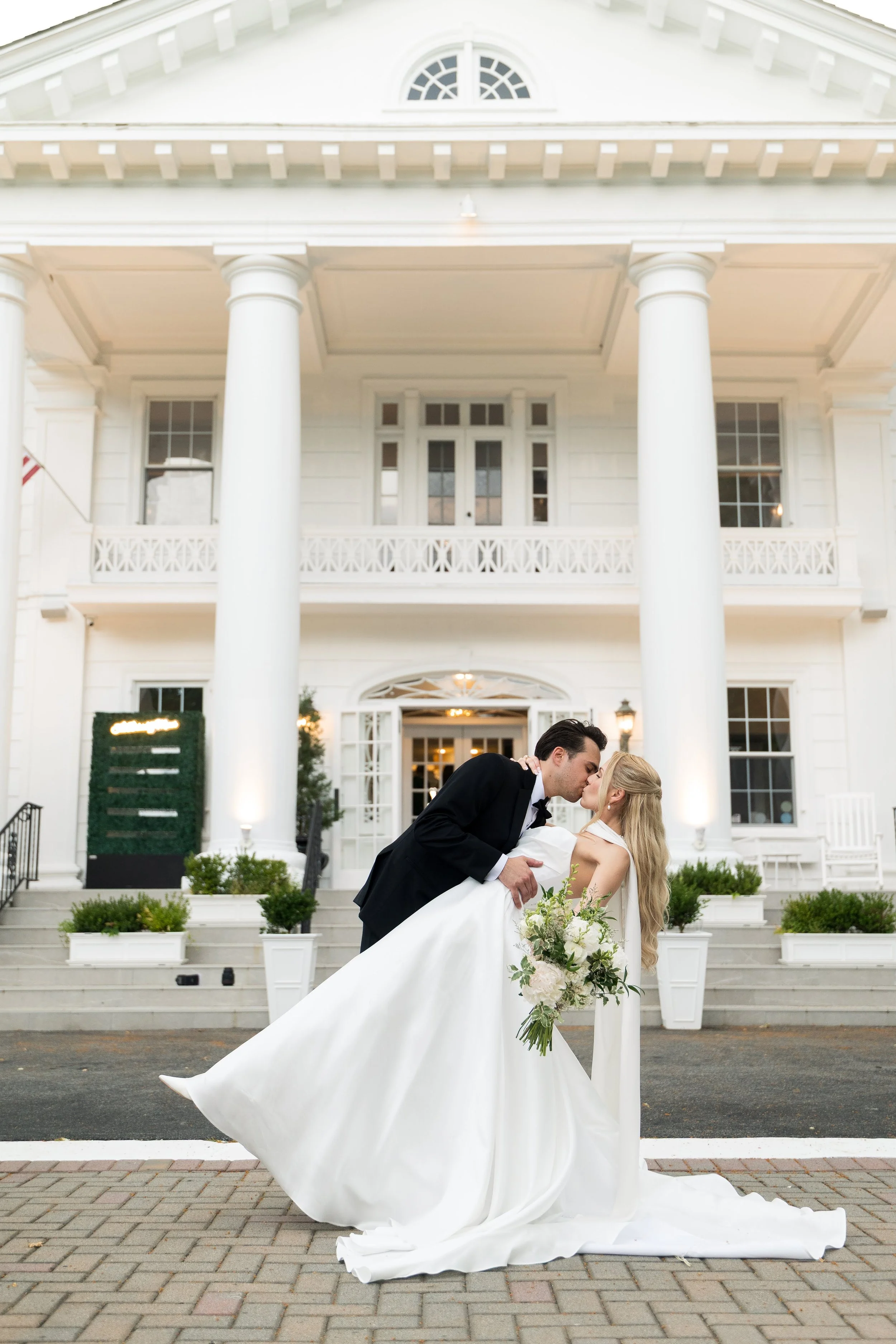 A newlywed couple sharing a kiss in front of a white, historic building with columns and large windows. The bride is holding a bouquet of white flowers and greenery, and the groom is dipping her as they kiss.