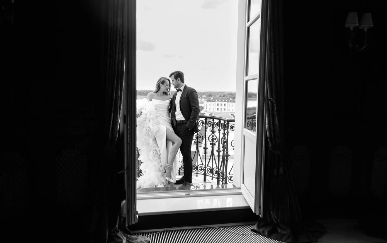 A bride and groom on a balcony, with the bride in a wedding dress and the groom in a tuxedo, looking at each other.