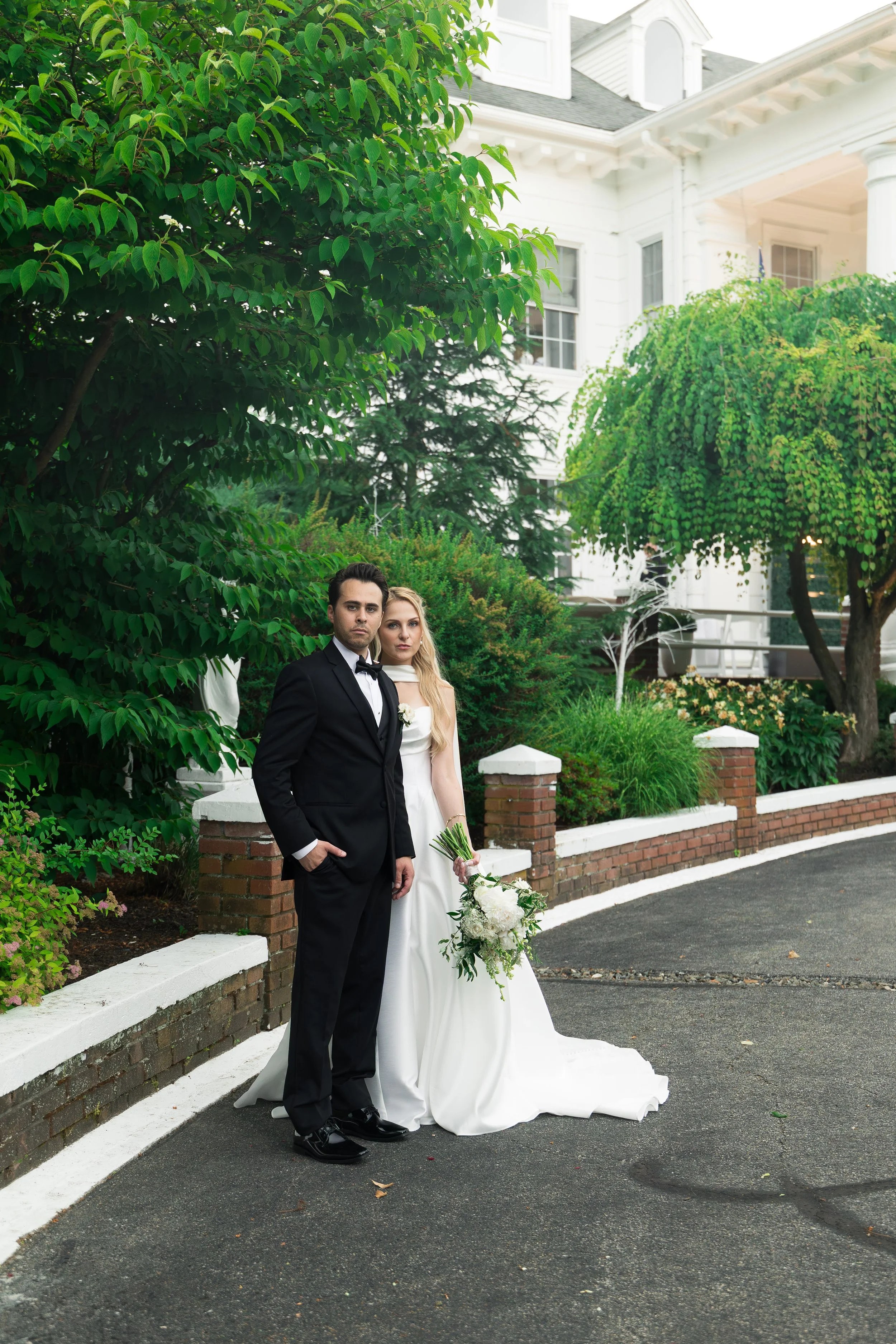 A bride and groom posing outdoors in front of a white house with green trees. The groom is wearing a black tuxedo, and the bride is in a white wedding gown holding a bouquet of white flowers.