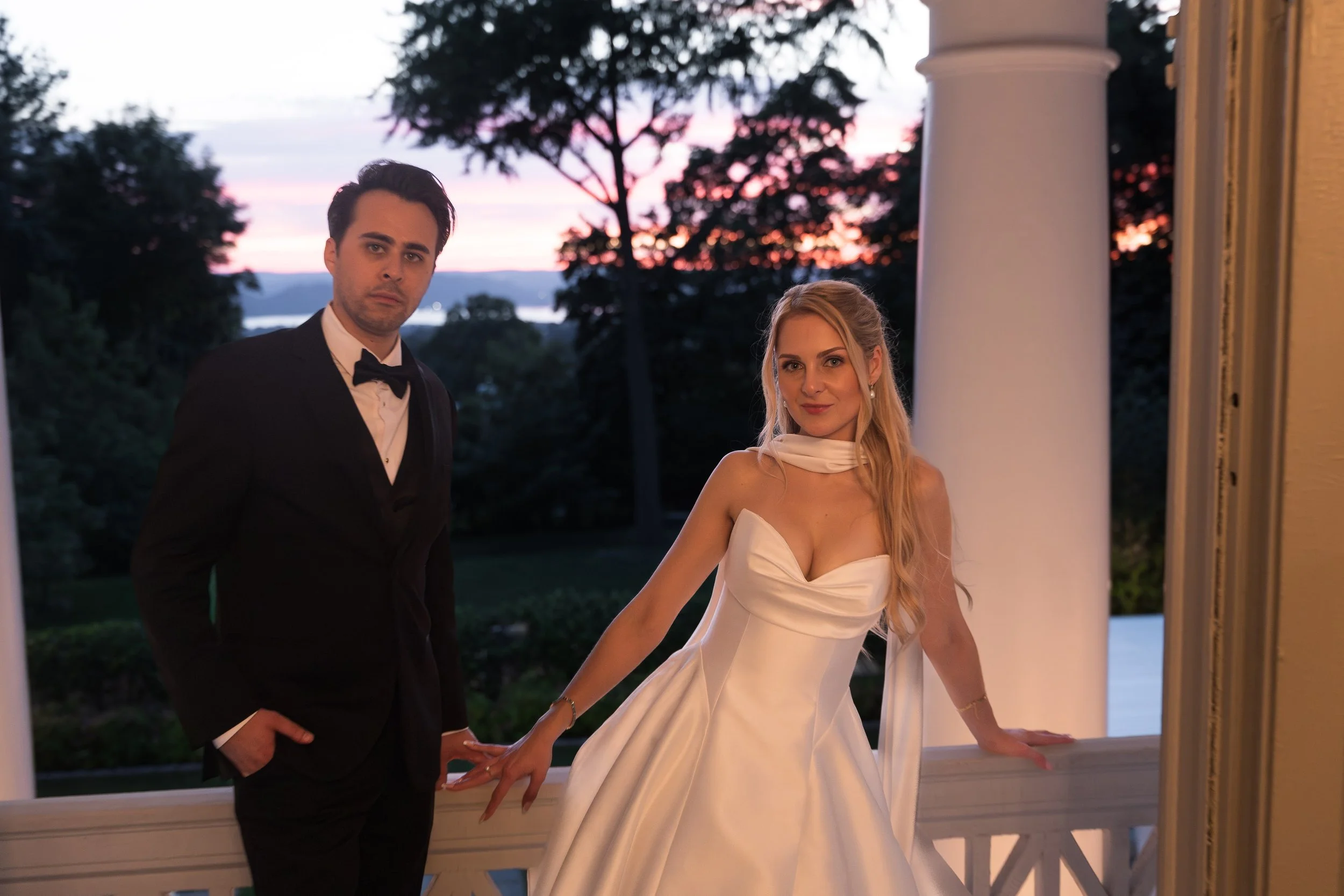 A man in a black tuxedo and a woman in a white wedding gown holding hands on a porch at sunset.