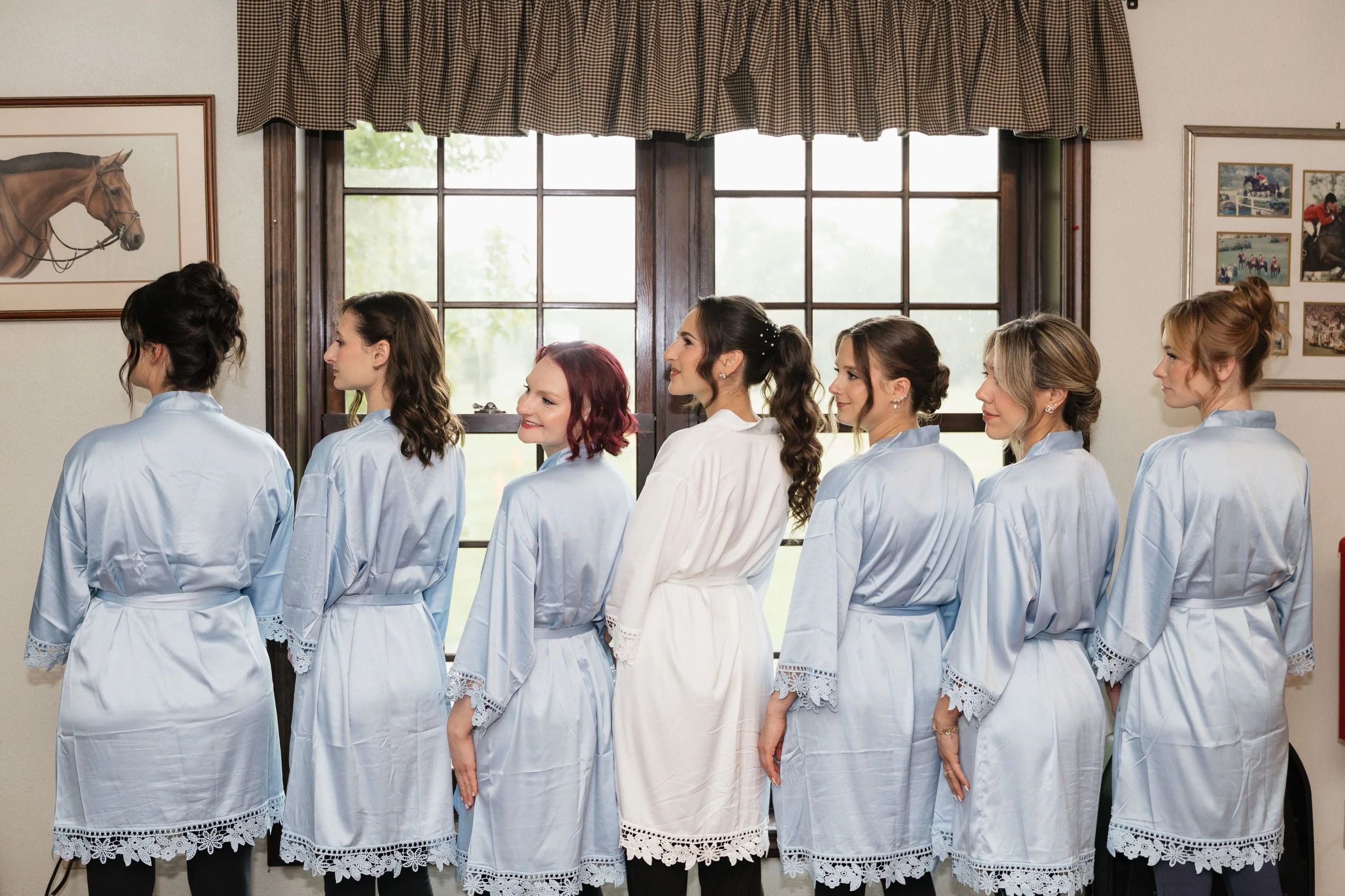 A group of women dressed in matching white robes, standing in a line, facing a window in a room decorated with pictures of horses.