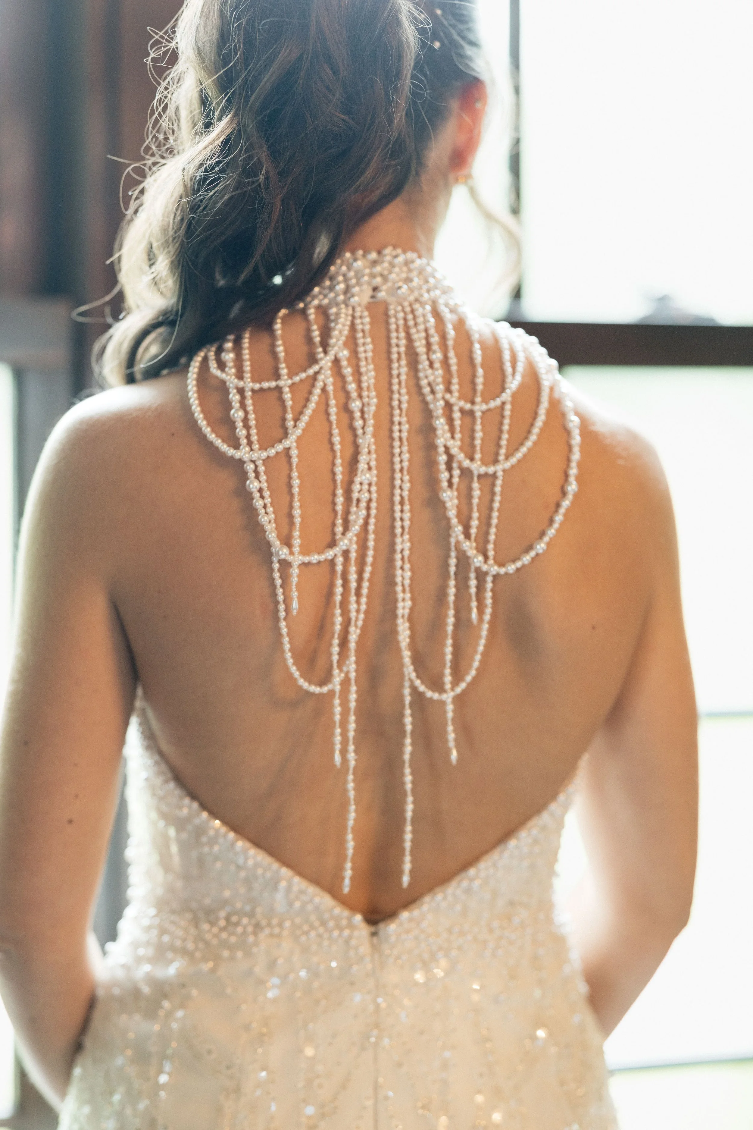 Back view of a woman in a wedding dress with pearl embellishments and a pearl necklace spread across her back, standing near a window with sunlight.