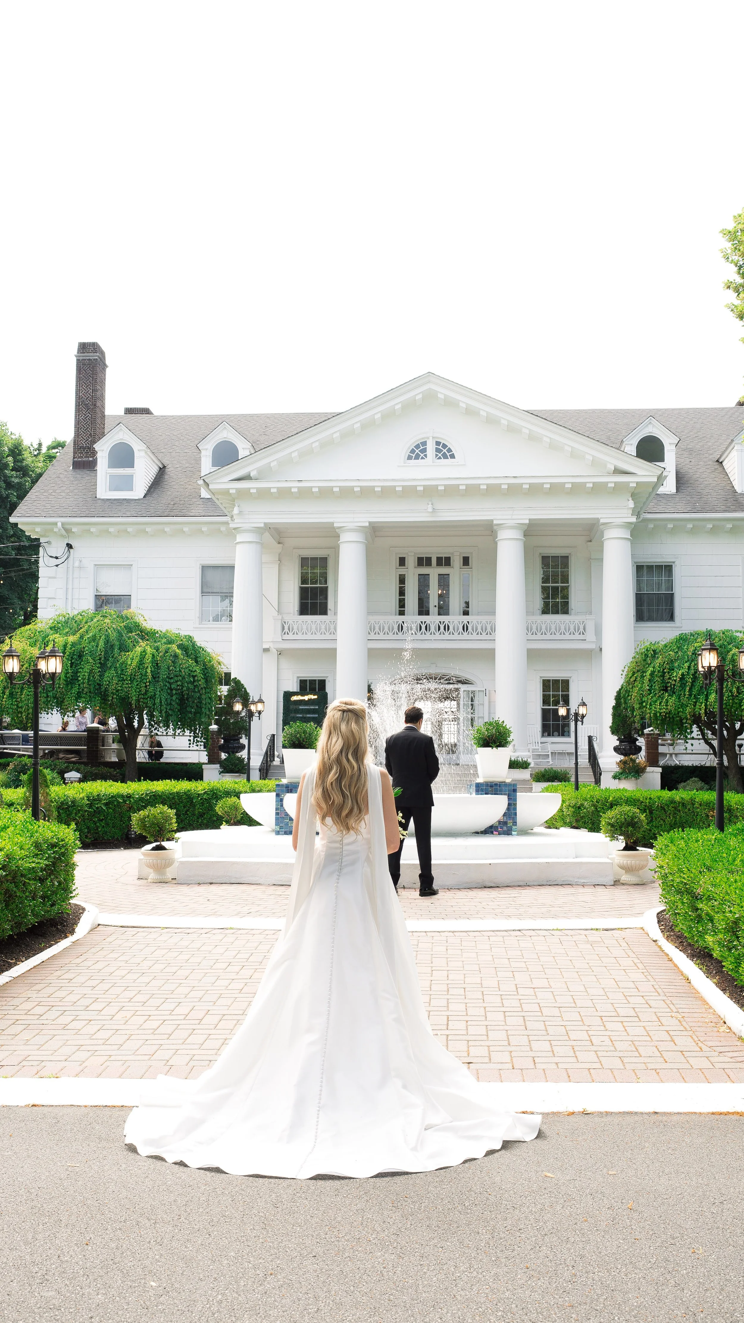 Bride and groom standing in front of a large white house with columns, fountain, and garden, during a wedding.