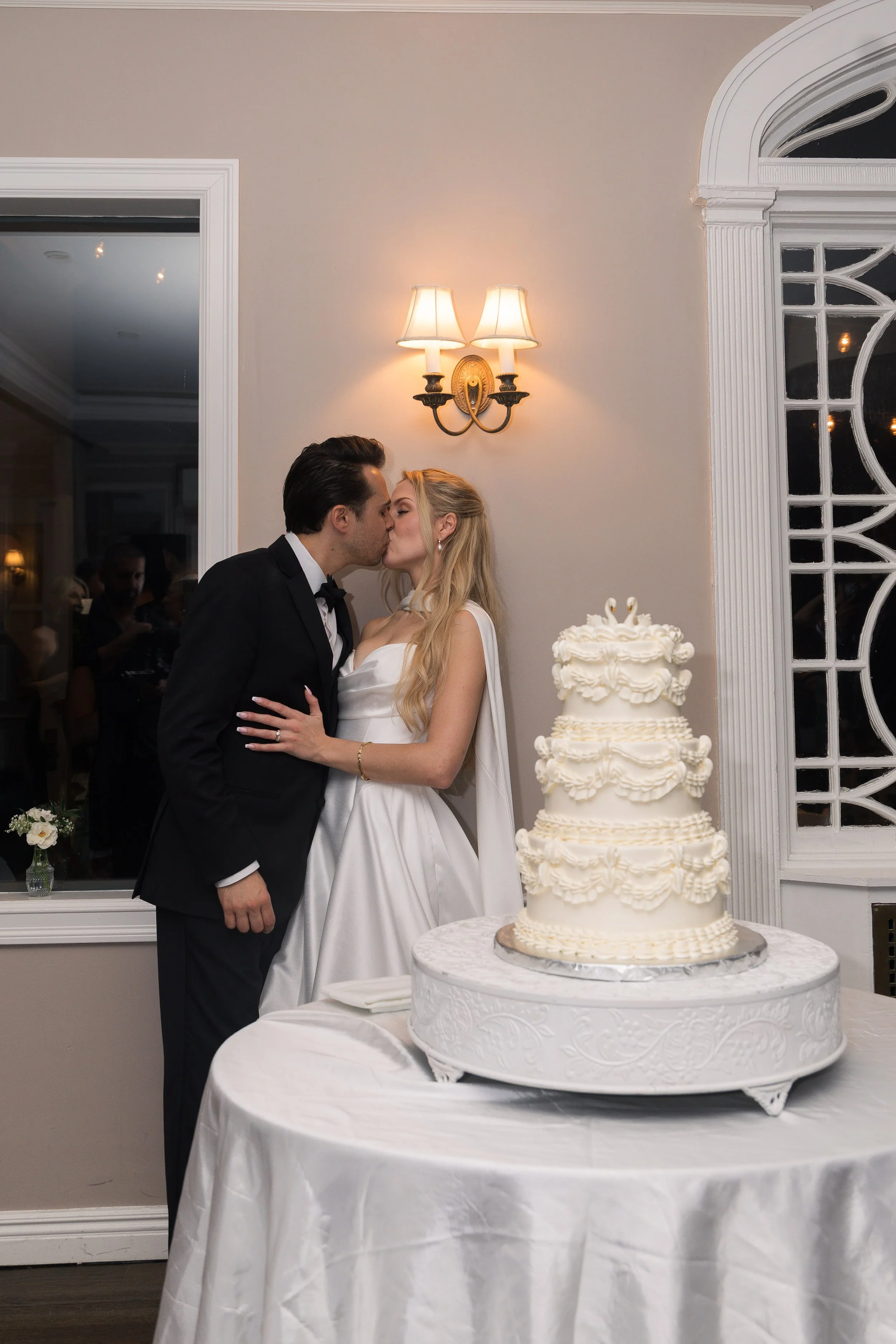 A bride and groom share a kiss at their wedding reception, with a three-tiered wedding cake on a table in front of them.
