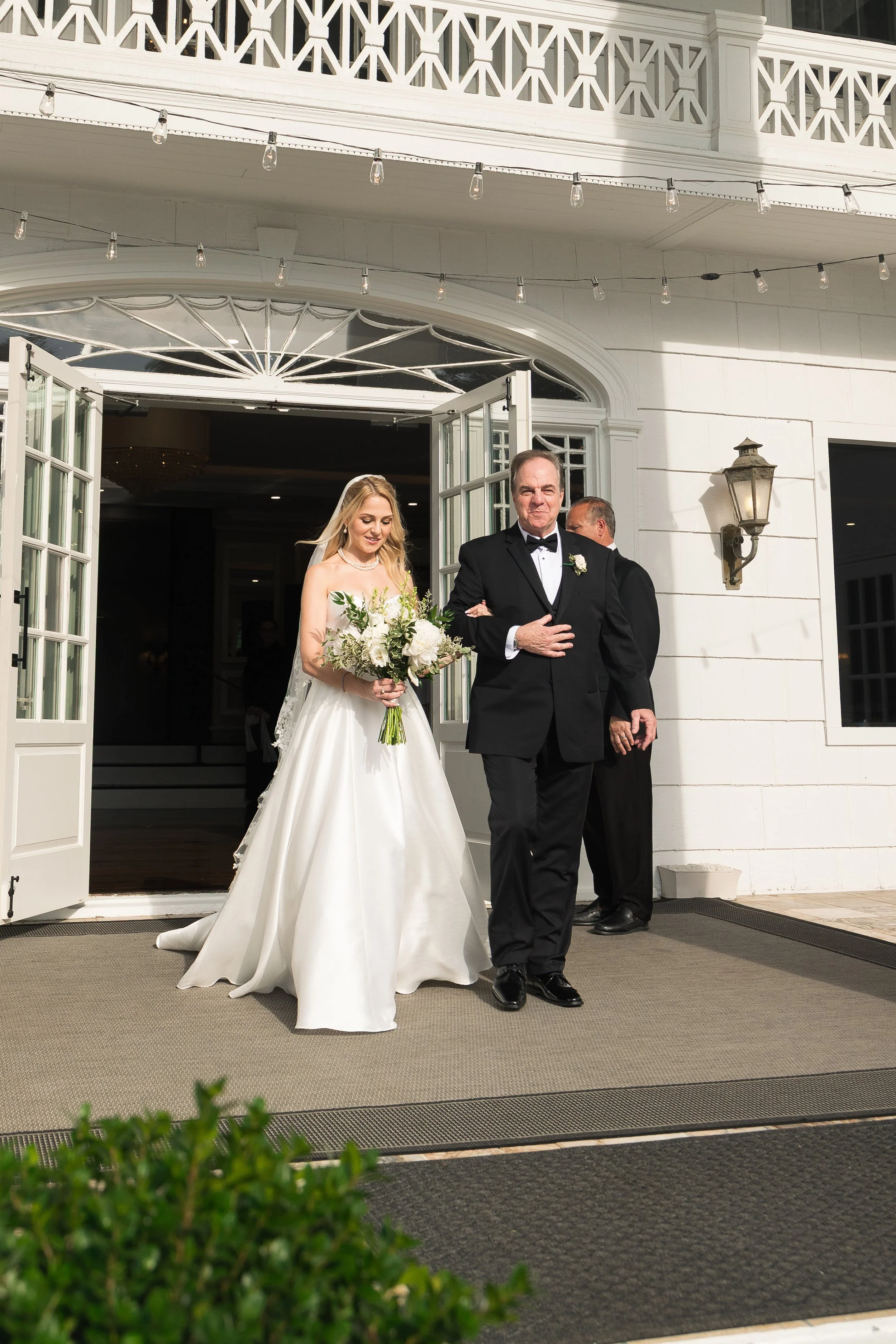 A bride in a white wedding dress with a bouquet of white and green flowers walks out of a building, accompanied by an older man in a tuxedo with a boutonnière, as part of a wedding procession.