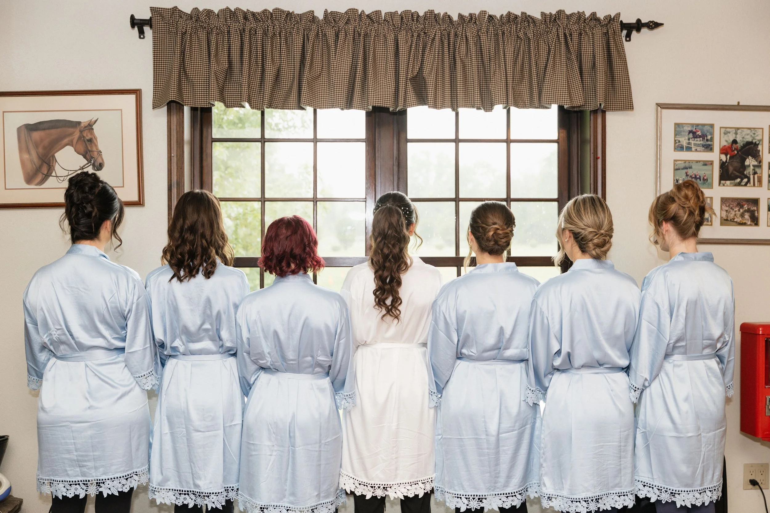 Seven women in white robes standing in front of a large window with curtains, facing away from the camera, with framed pictures of horse racing on the walls.
