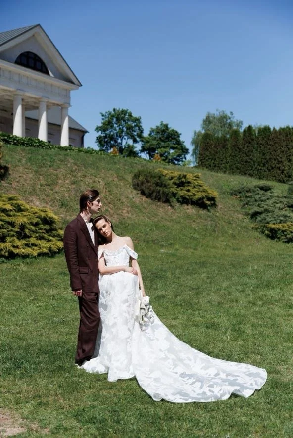 A couple dressed in wedding attire standing on a grassy hill outside a large white house with columns, with trees and bushes in the background.