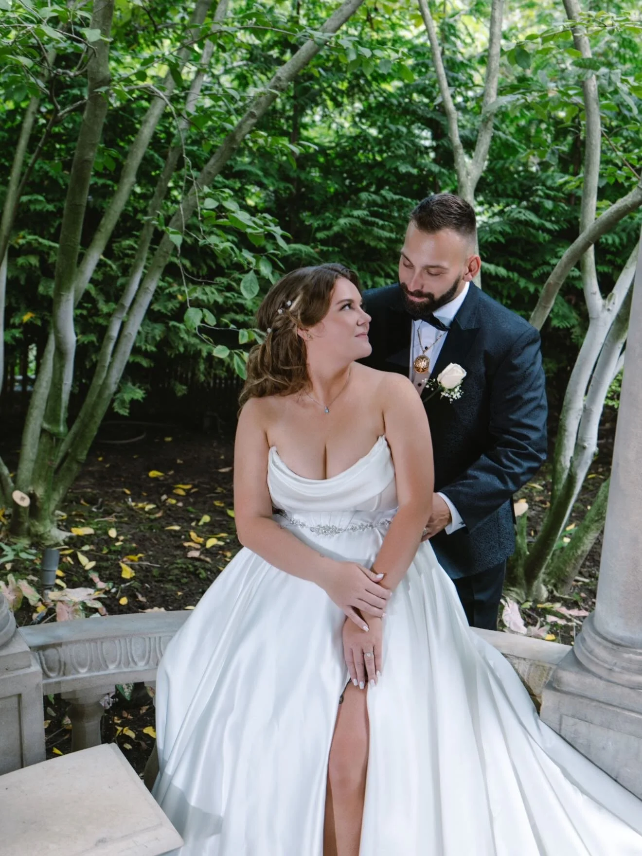 A bride and groom posing outdoors in a garden with lush green trees. She is sitting on a stone bench, wearing a white wedding gown, and he is standing behind her in a black tuxedo with a boutonniere, gazing at her affectionately.