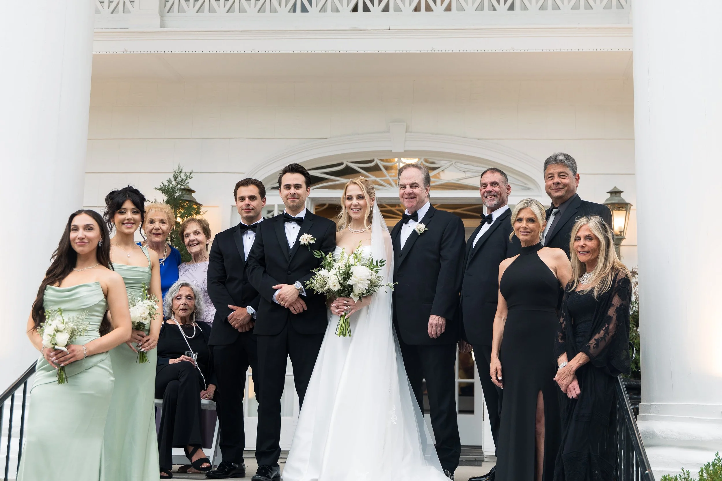 A group of wedding guests and the bride and groom standing outside a white building with columns. The bride holds a bouquet and wears a white wedding dress. The groom wears a black tuxedo. Women in dresses and men in tuxedos are smiling for the photo