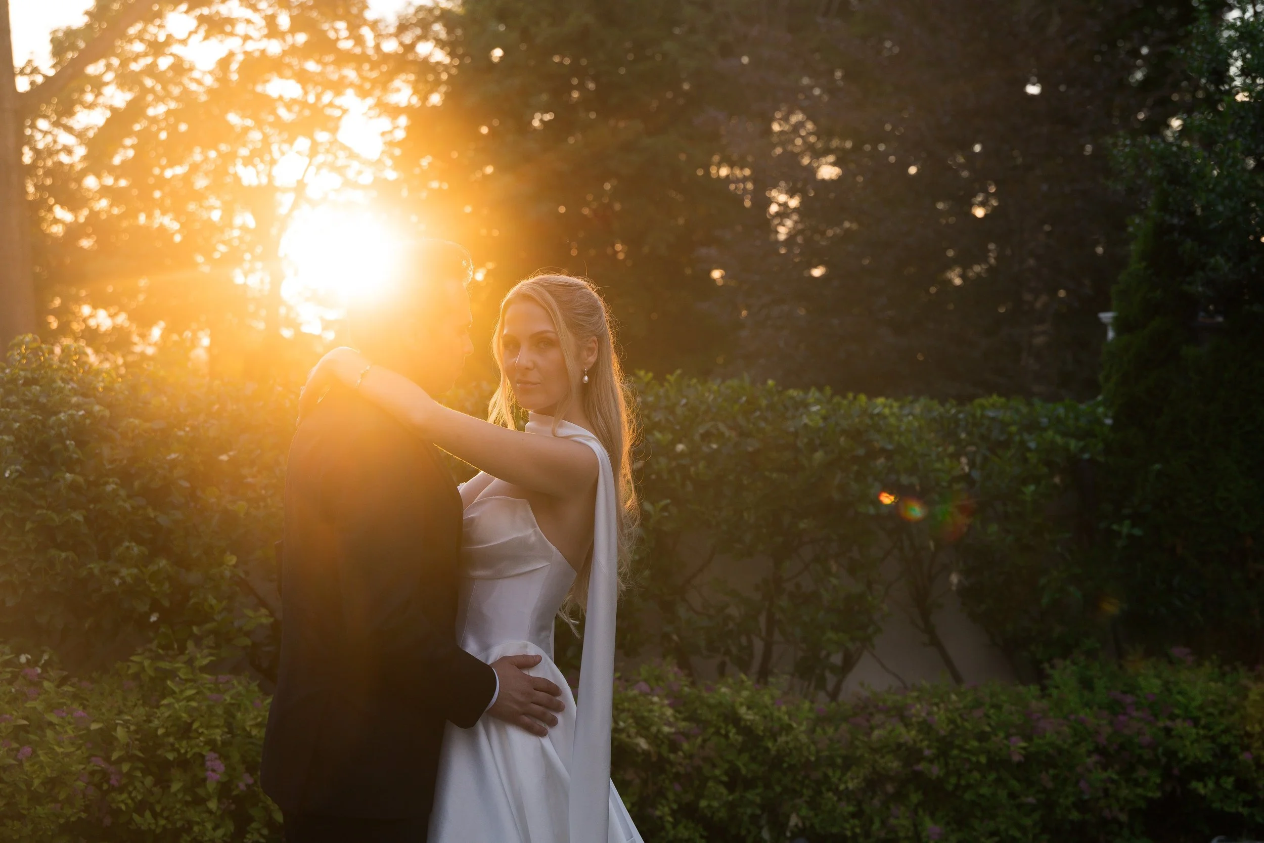 A couple dressed in wedding attire, the man in a black suit and the woman in a white wedding dress, embrace outdoors during sunset with sun filtering through trees in the background.