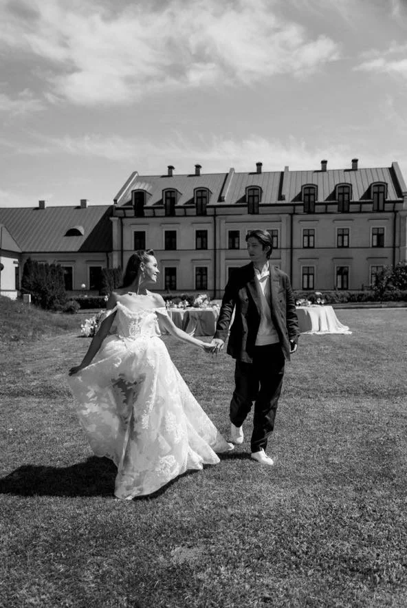 A bride and groom holding hands and dancing outdoors on a grassy lawn with a large historic building in the background, under a partly cloudy sky.