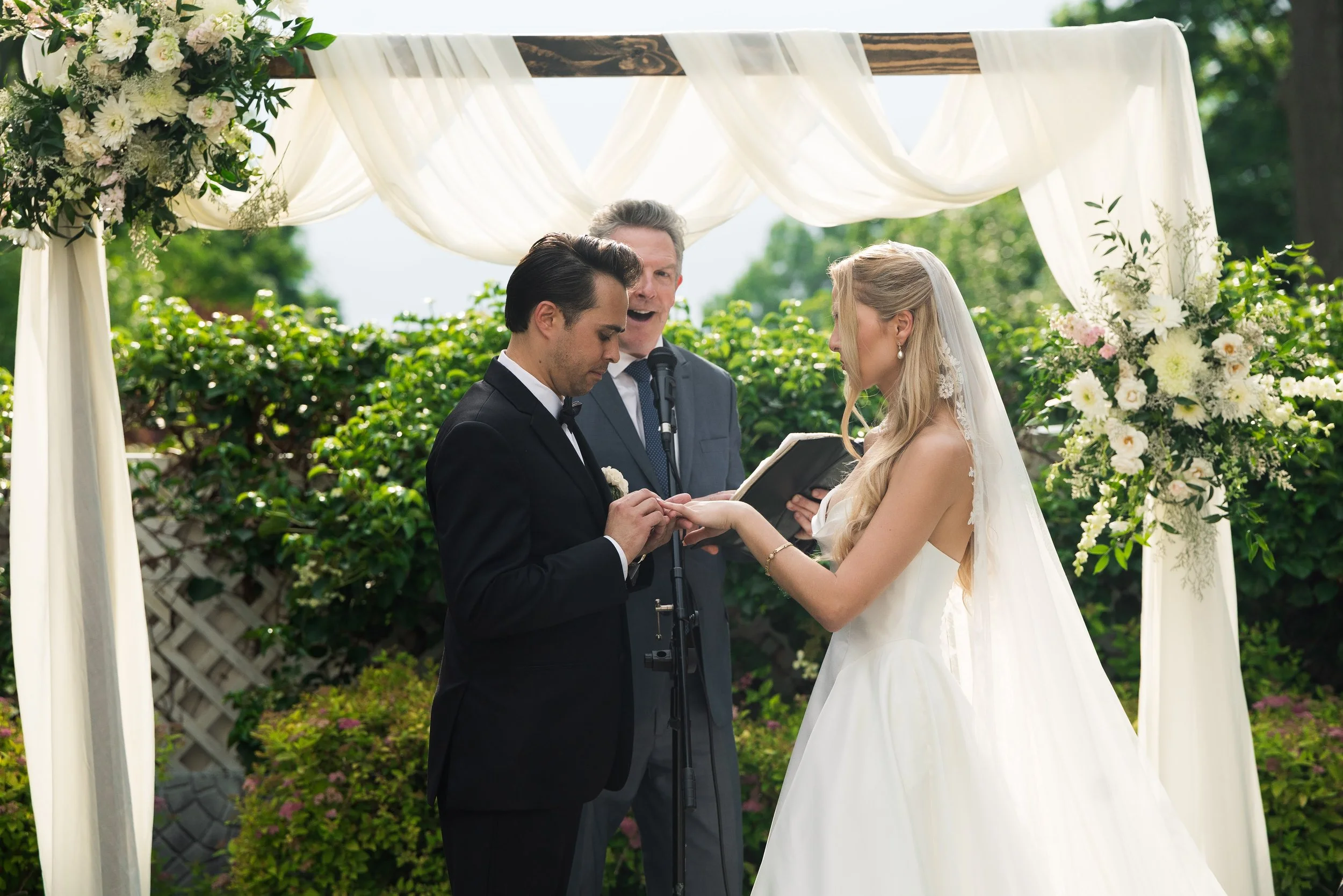 A couple getting married outdoors under a decorated canopy with white fabric and flowers, with an officiant standing between them.