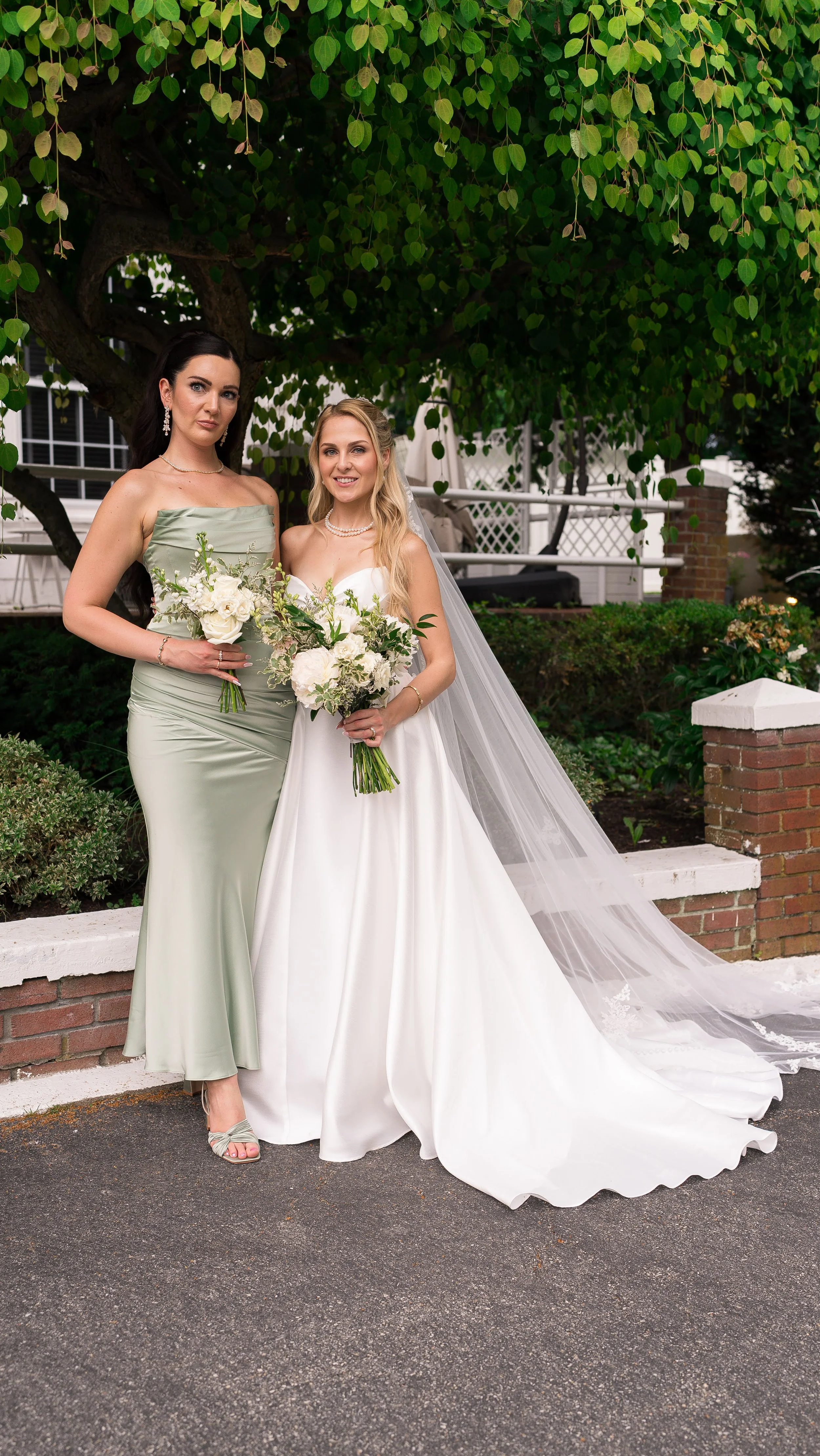 Two women in formal attire standing outdoors under a leafy tree, holding bouquets of white flowers, one in a satin gown with a tulle veil, and the other in a green dress with jewelry.