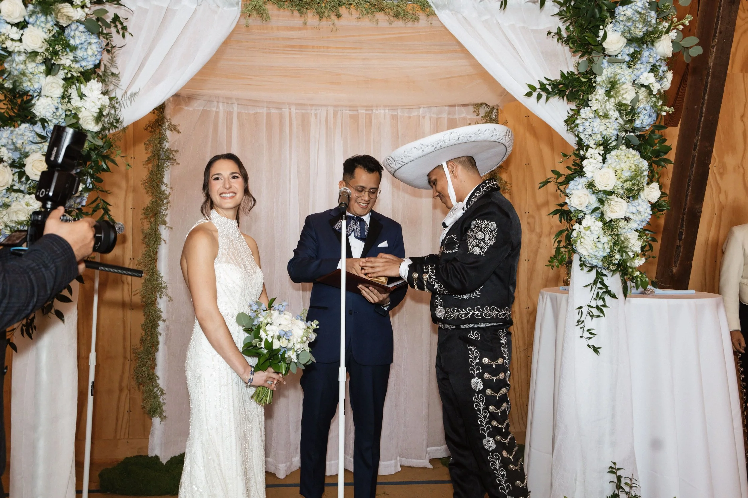 A couple getting married in a wedding ceremony, with two officiants, under an arch decorated with white and blue flowers and green foliage.