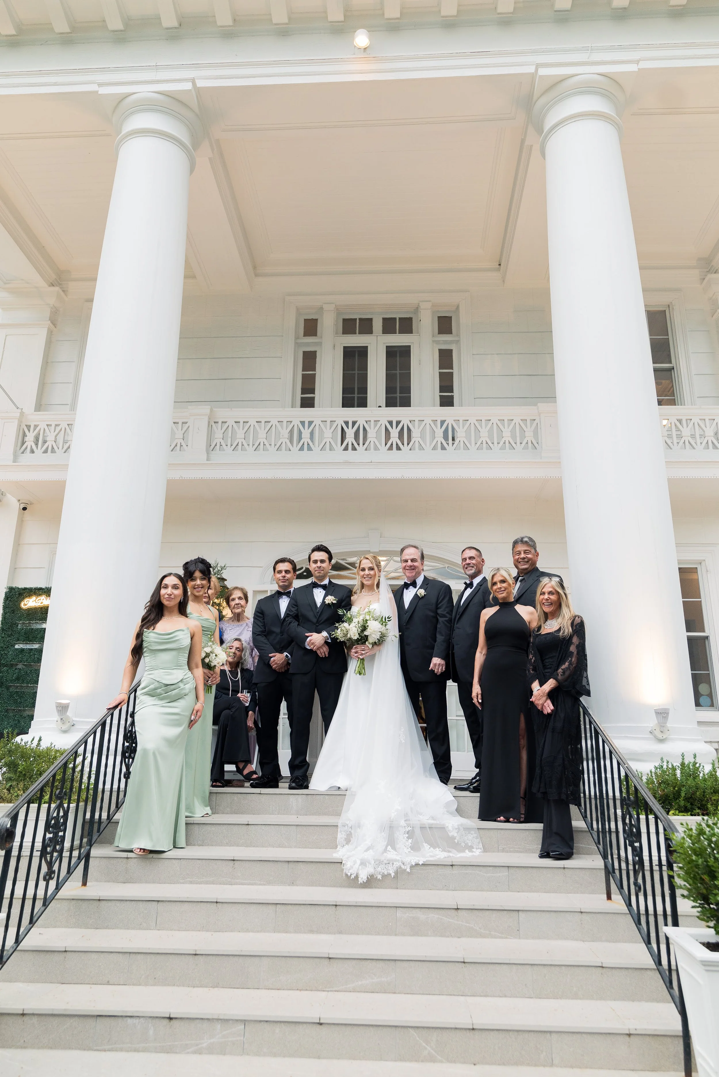 A wedding group photo on the steps of a white classical building with large columns. The bride in a white wedding dress and the groom in a tuxedo are at the center, surrounded by family and friends dressed in elegant attire.