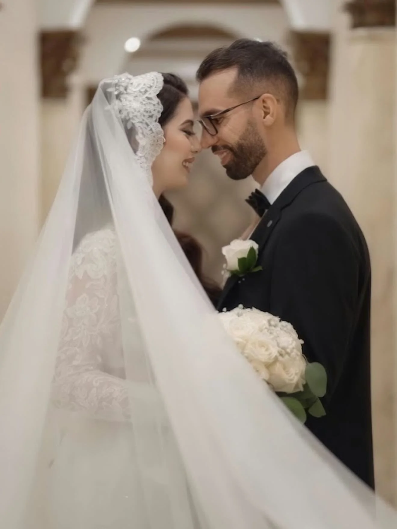 A bride and groom smiling and touching foreheads in a wedding ceremony. The bride is wearing a lace wedding dress and a veil, and holding a bouquet of white roses. The groom is dressed in a tuxedo with a bow tie and glasses.