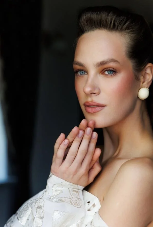 A young woman with blue eyes, fair skin, and dark brown hair styled back, wearing pearl earrings and an off-the-shoulder white lace top, looking thoughtfully at the camera with her hand near her face.