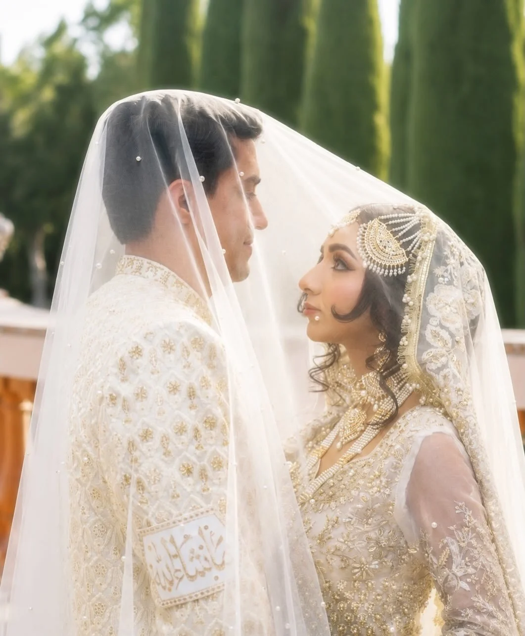 A bride and groom face each other under a wedding veil, dressed in traditional white and gold attire with ornate jewelry, outdoors with green trees in the background.