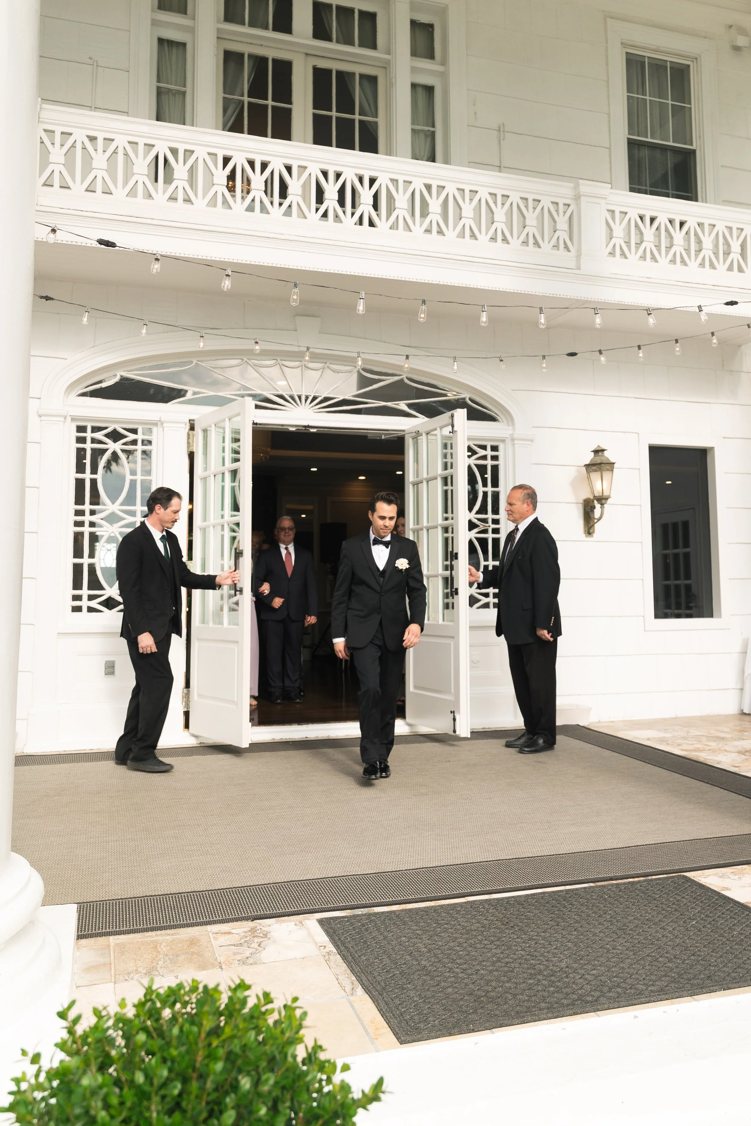 A man in a tuxedo walking out of a white building with open doors, flanked by two men in suits, with others inside the building. The building has a balcony, string lights above the doorway, and a decorative lantern on the wall.