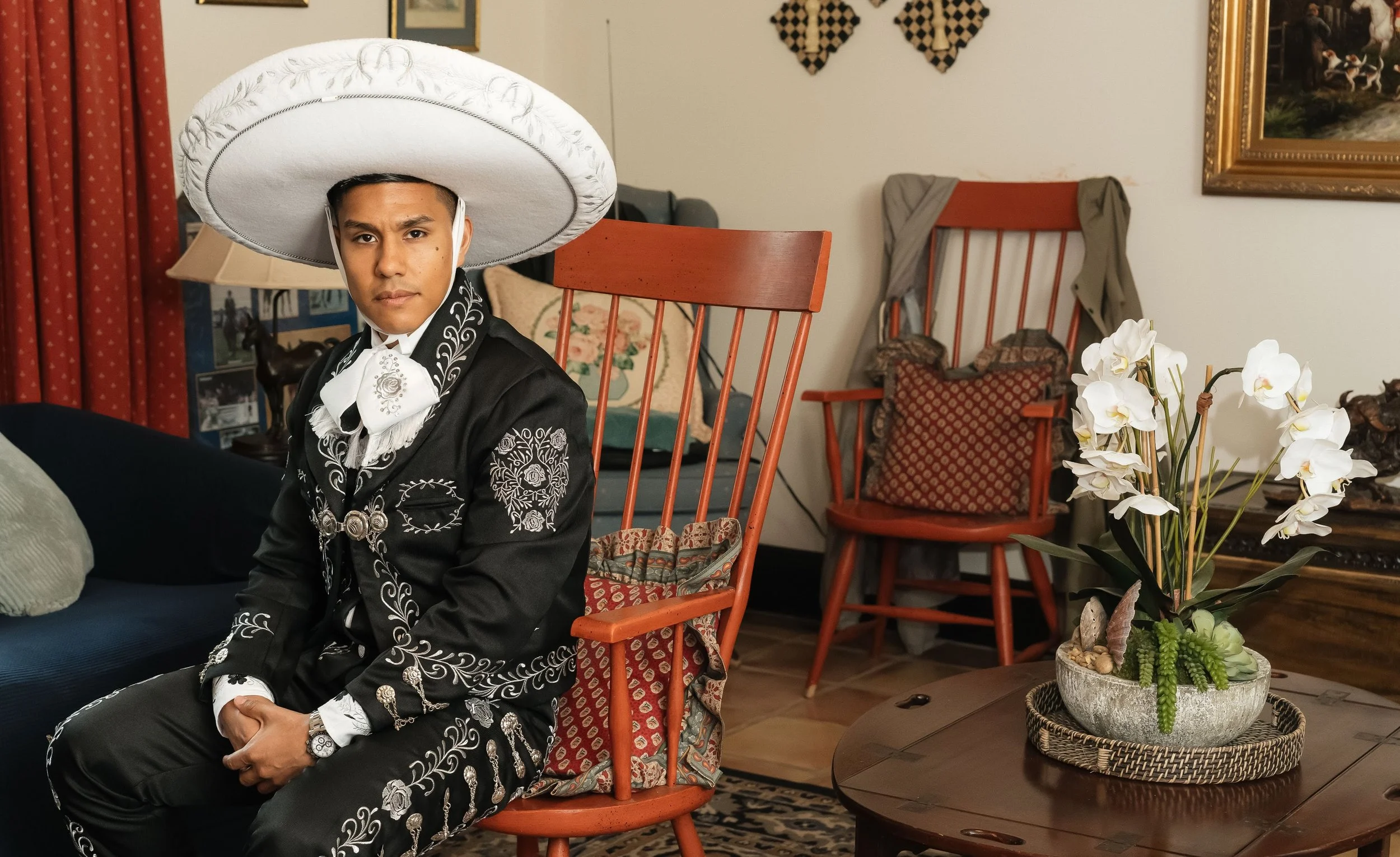 A man wearing traditional Mexican mariachi attire, including a large sombrero, is sitting in a living room with wooden furniture and decorative pillows. There are flower arrangements on a table to the right.