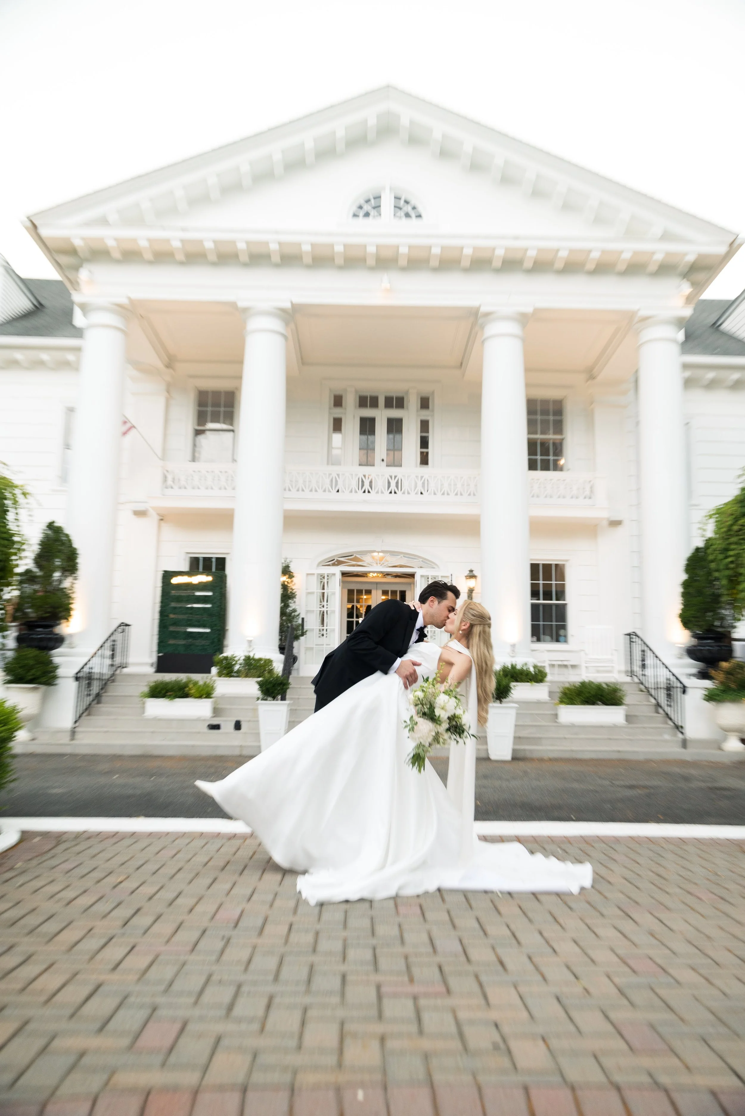 A bride and groom kissing in front of a large white house with columns, during a wedding celebration.