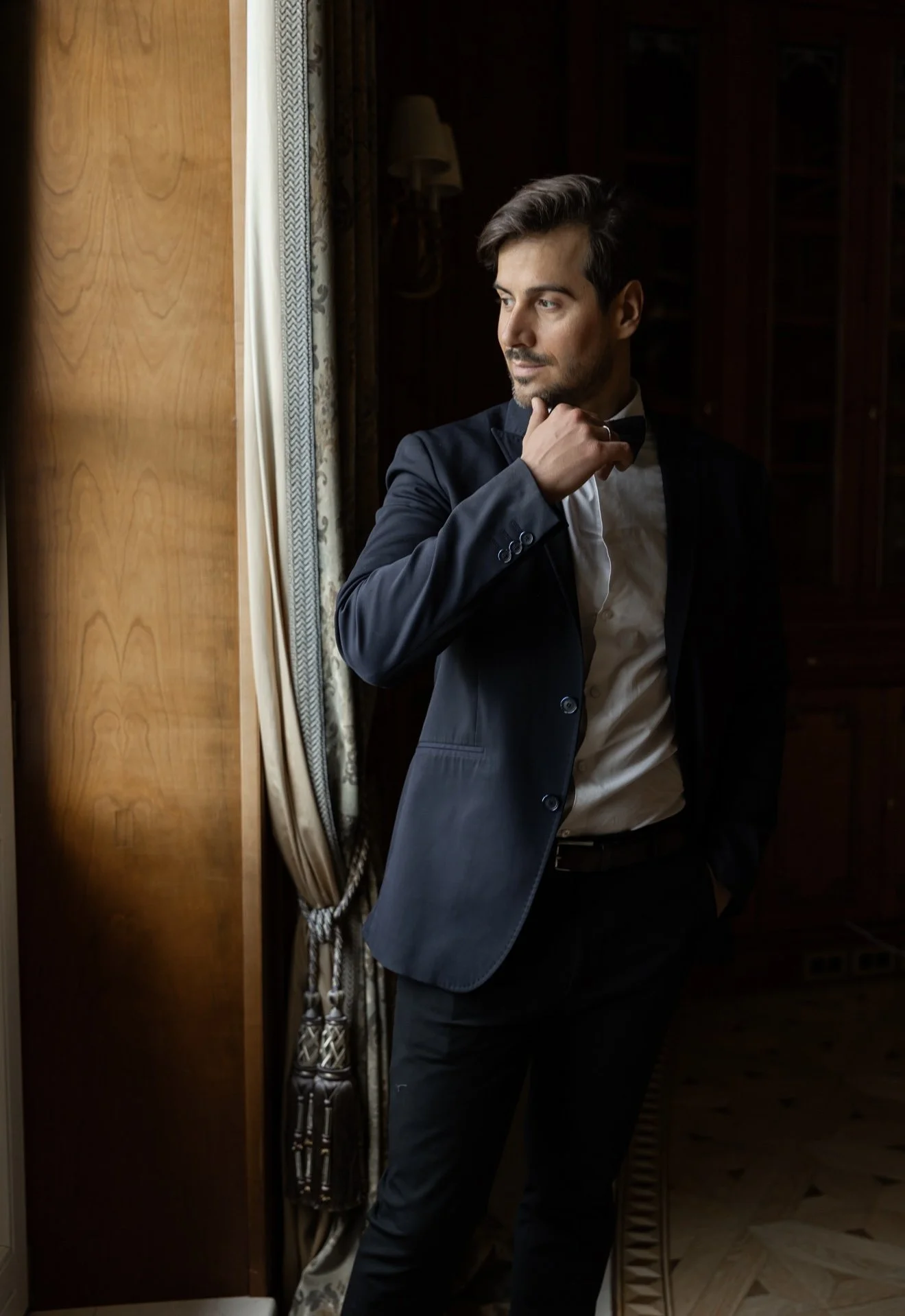 A man dressed in formal attire, a suit and bowtie, standing indoors near a window with patterned curtains, adjusting his bowtie and looking thoughtfully outside.