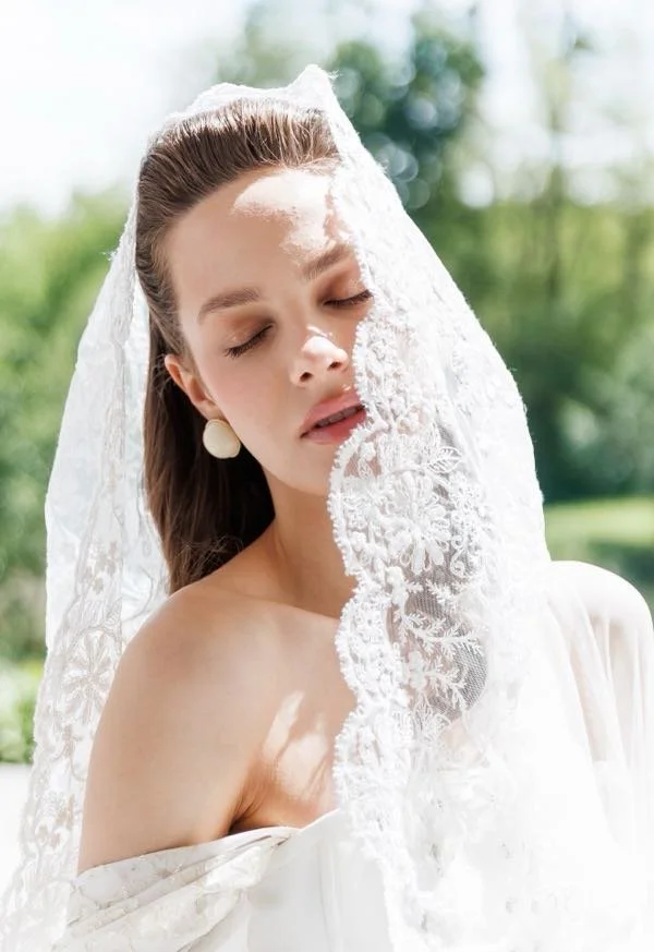 A woman with long brown hair, eyes closed, partially covered with a delicate lace veil outdoors on a sunny day.