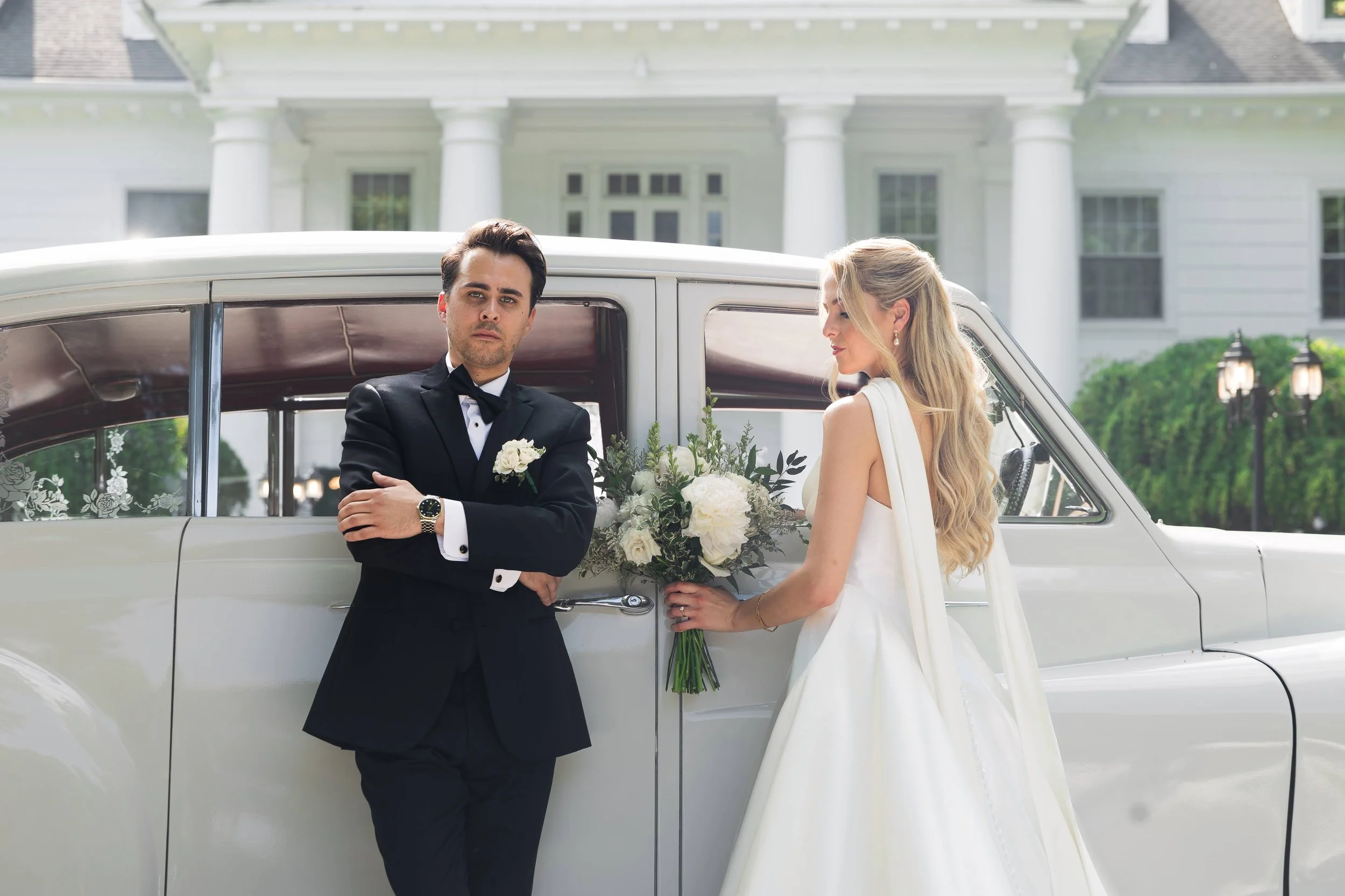 A groom in a black tuxedo stands with arms crossed, looking serious, next to a bride in a white wedding dress holding a bouquet, in front of a vintage car on the driveway of a large white house.