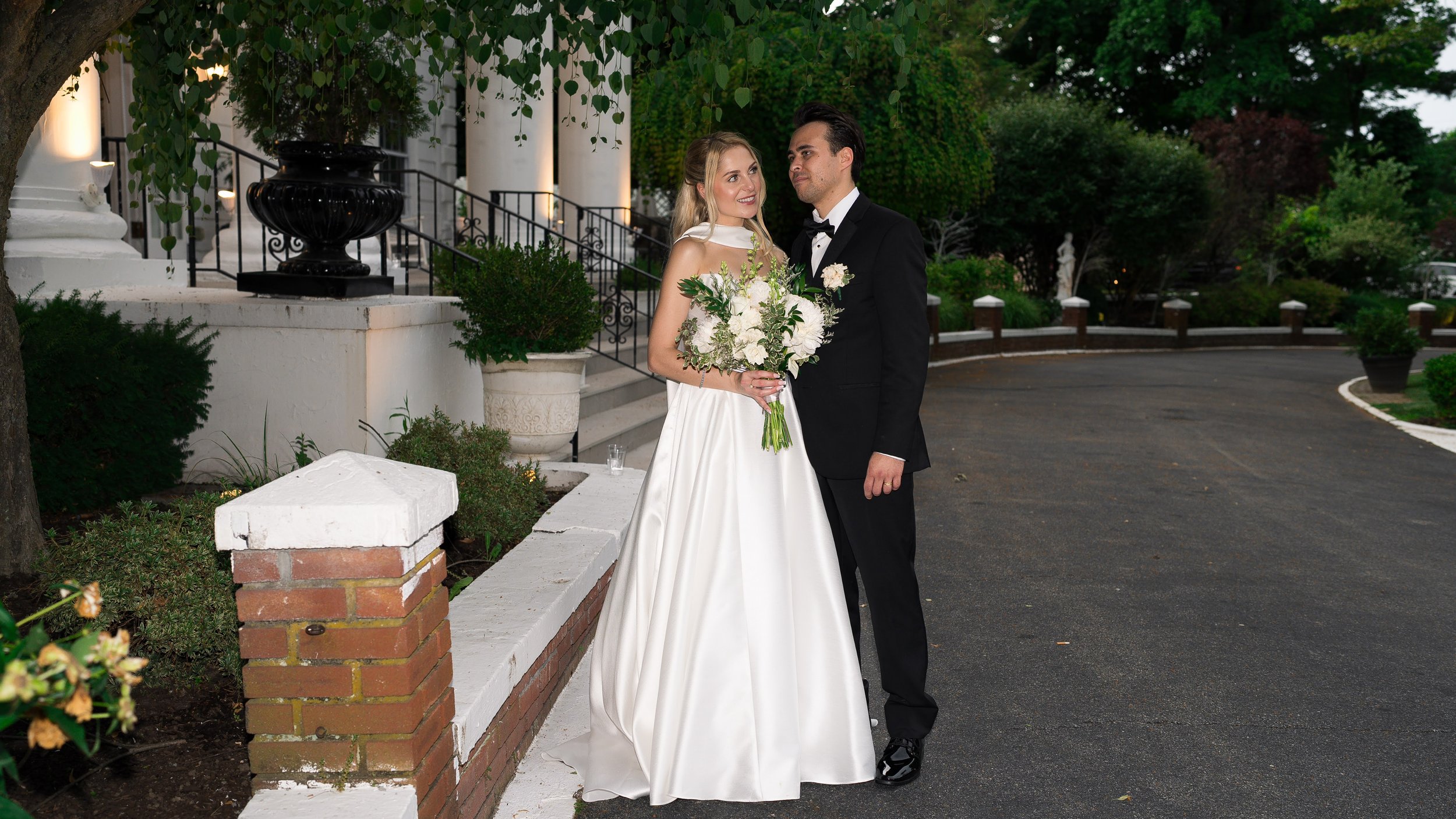 A bride and groom stand outside a building at sunset, with the bride holding a bouquet of white flowers and greenery, both dressed in wedding attire.