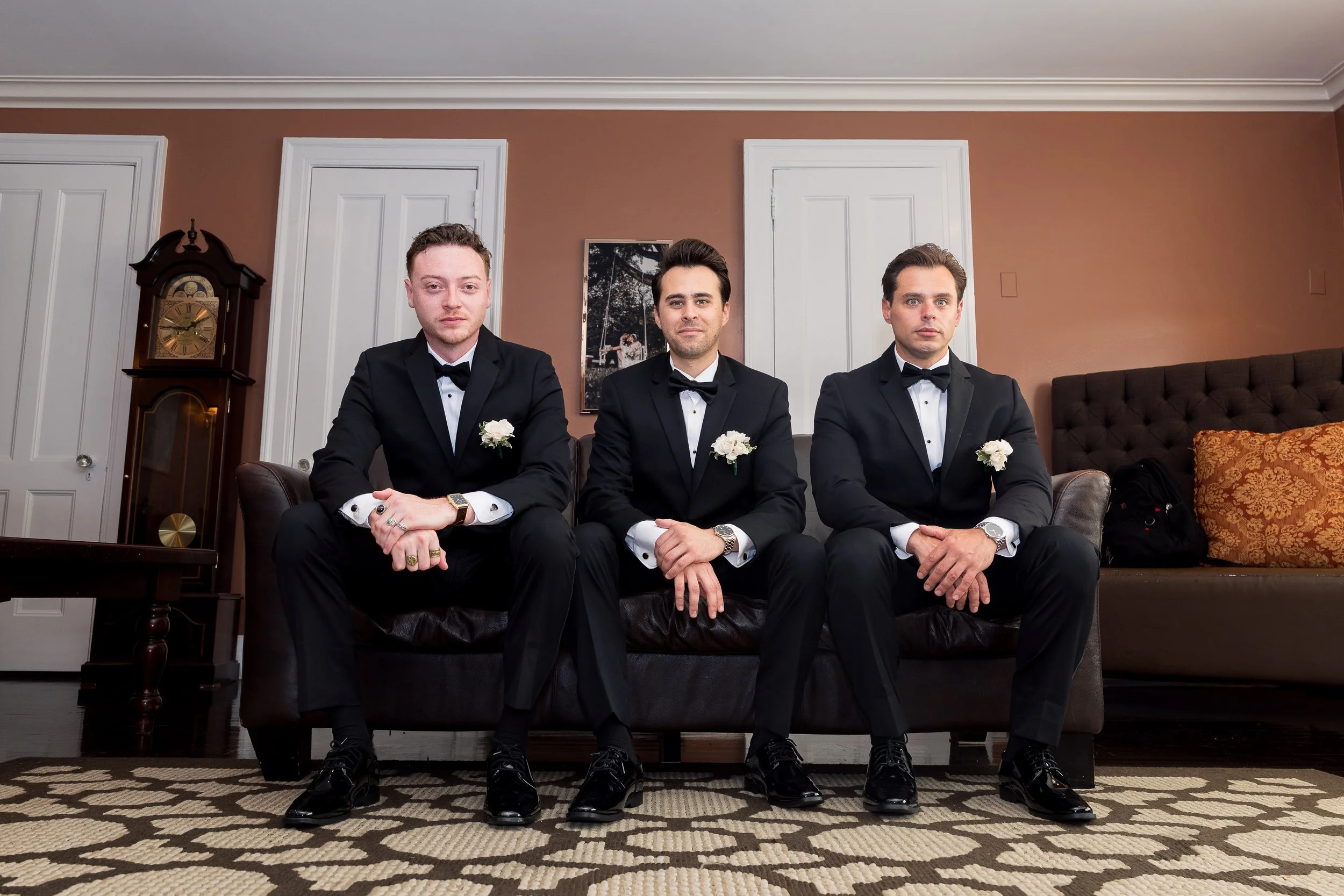 Three men in tuxedos with white shirts and black bow ties, sitting on a dark leather sofa. Each man is wearing a white boutonniere on his left lapel, and watches on their left wrists. The room has brown walls, a grandfather clock on the left, a paint