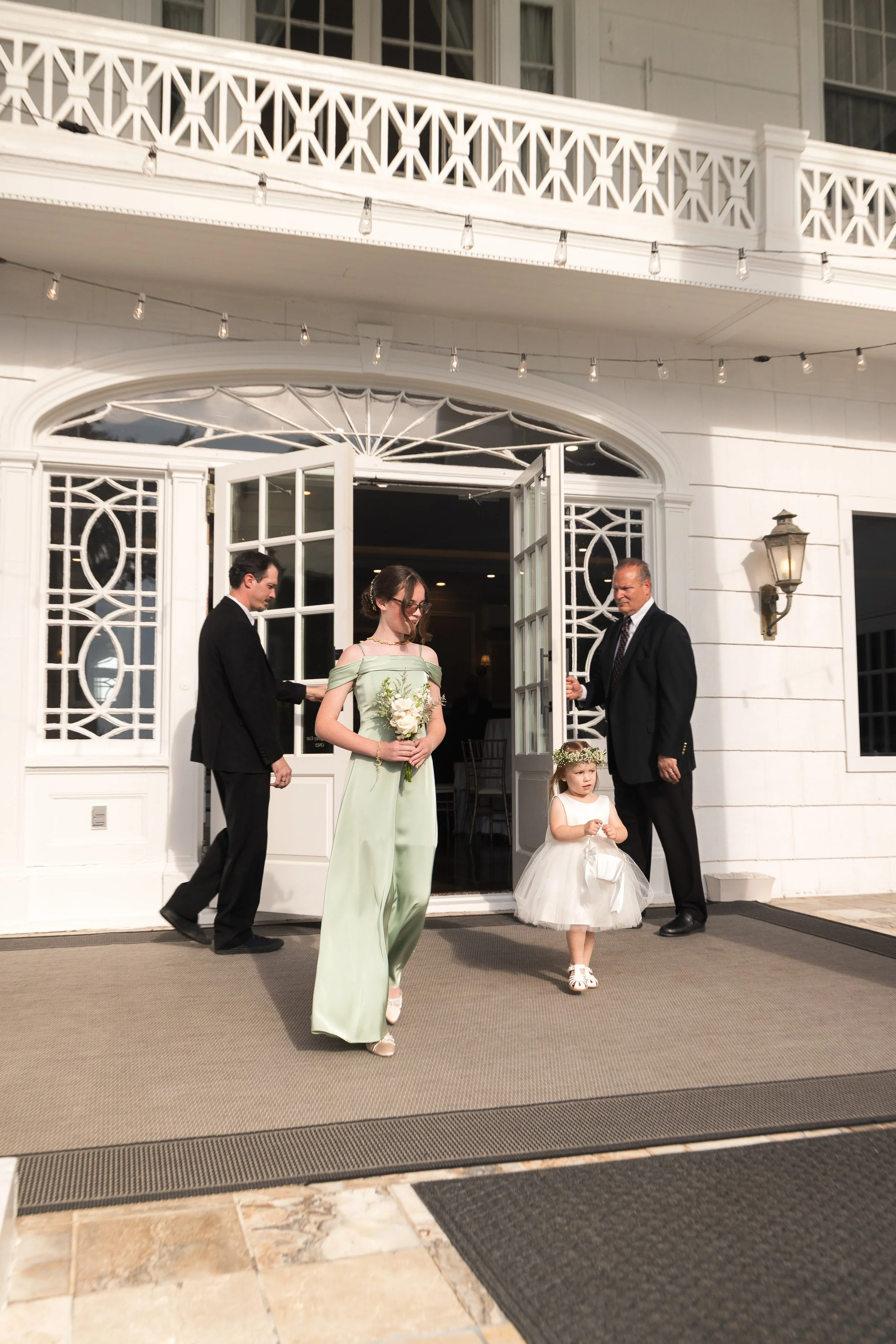 A wedding ceremony taking place outside a white building with open doors and decorative windows. Two men in suits and a woman in an elegant dress are standing, with a young girl in a white dress and flower crown in the foreground.