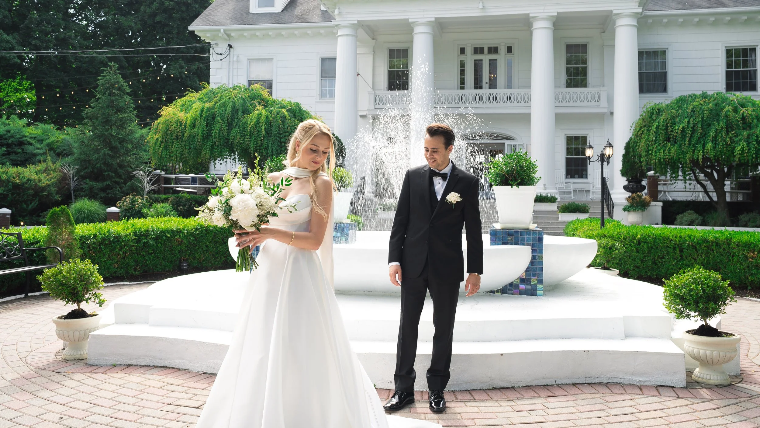 A bride and groom standing outdoors in front of a white house with columns. The bride is holding a bouquet of white flowers and is looking down, while the groom is smiling and looking at her.