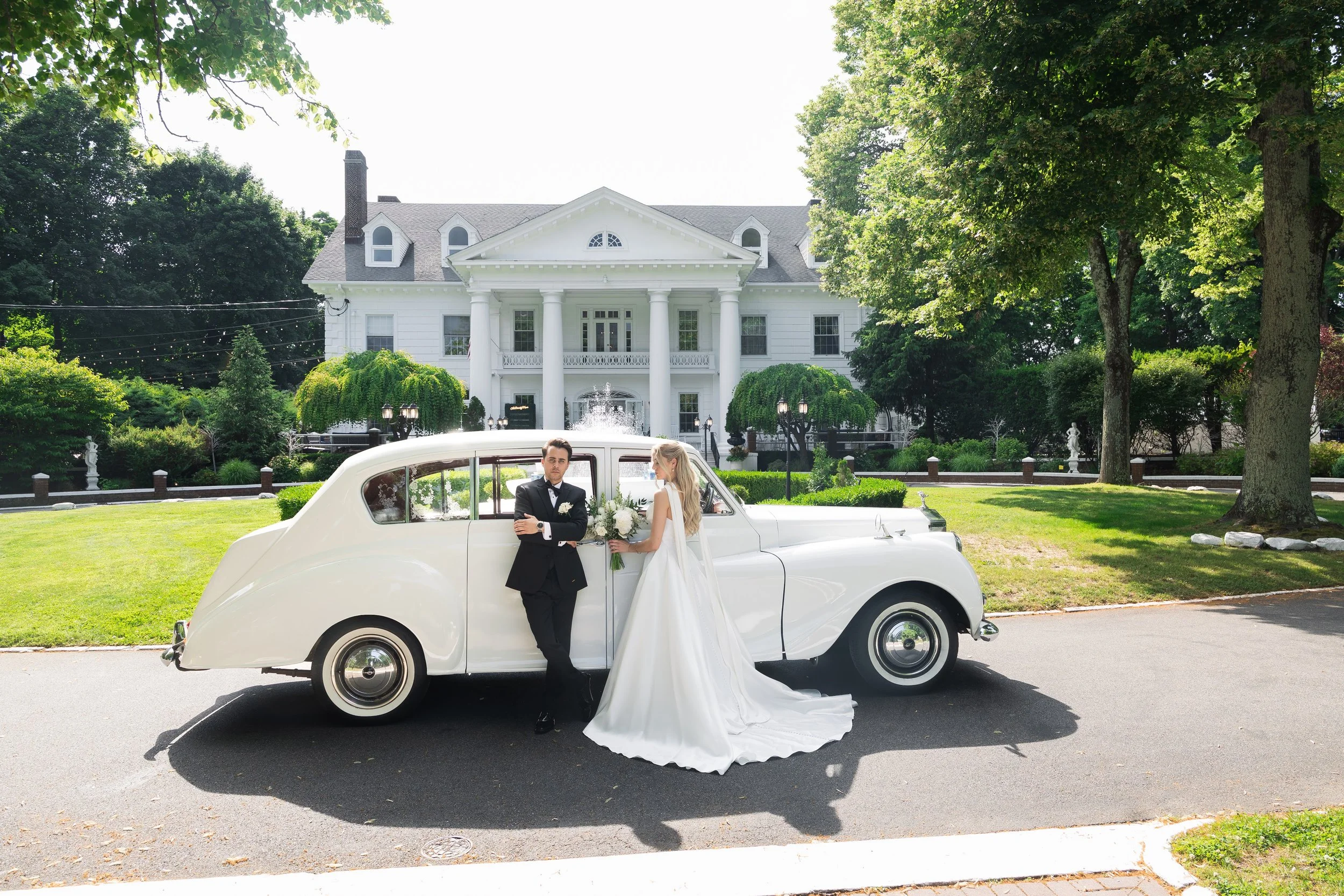 A bride and groom standing next to a vintage white car in front of a large white mansion, with trees and greenery surrounding the scene.