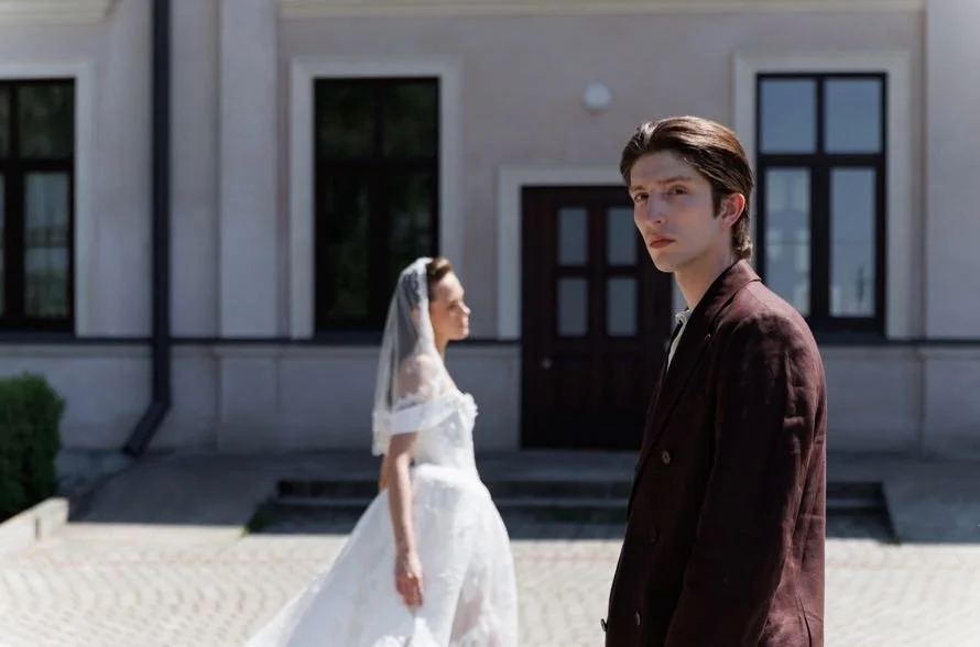 A young man in a brown suit stands outdoors in front of a building, looking at the camera. A woman in a white wedding dress with a veil is in the background, walking away from the building.
