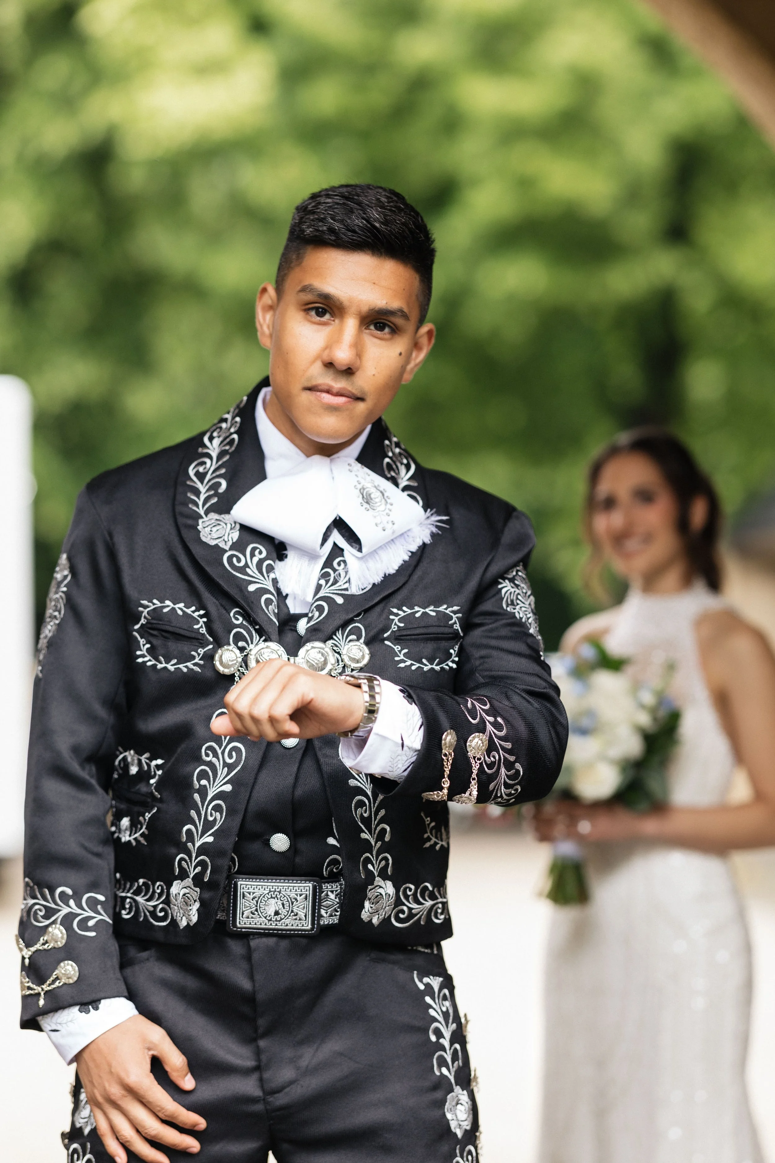A man dressed in traditional Mexican charro attire standing outdoors with a woman in the background holding a bouquet of flowers, smiling.