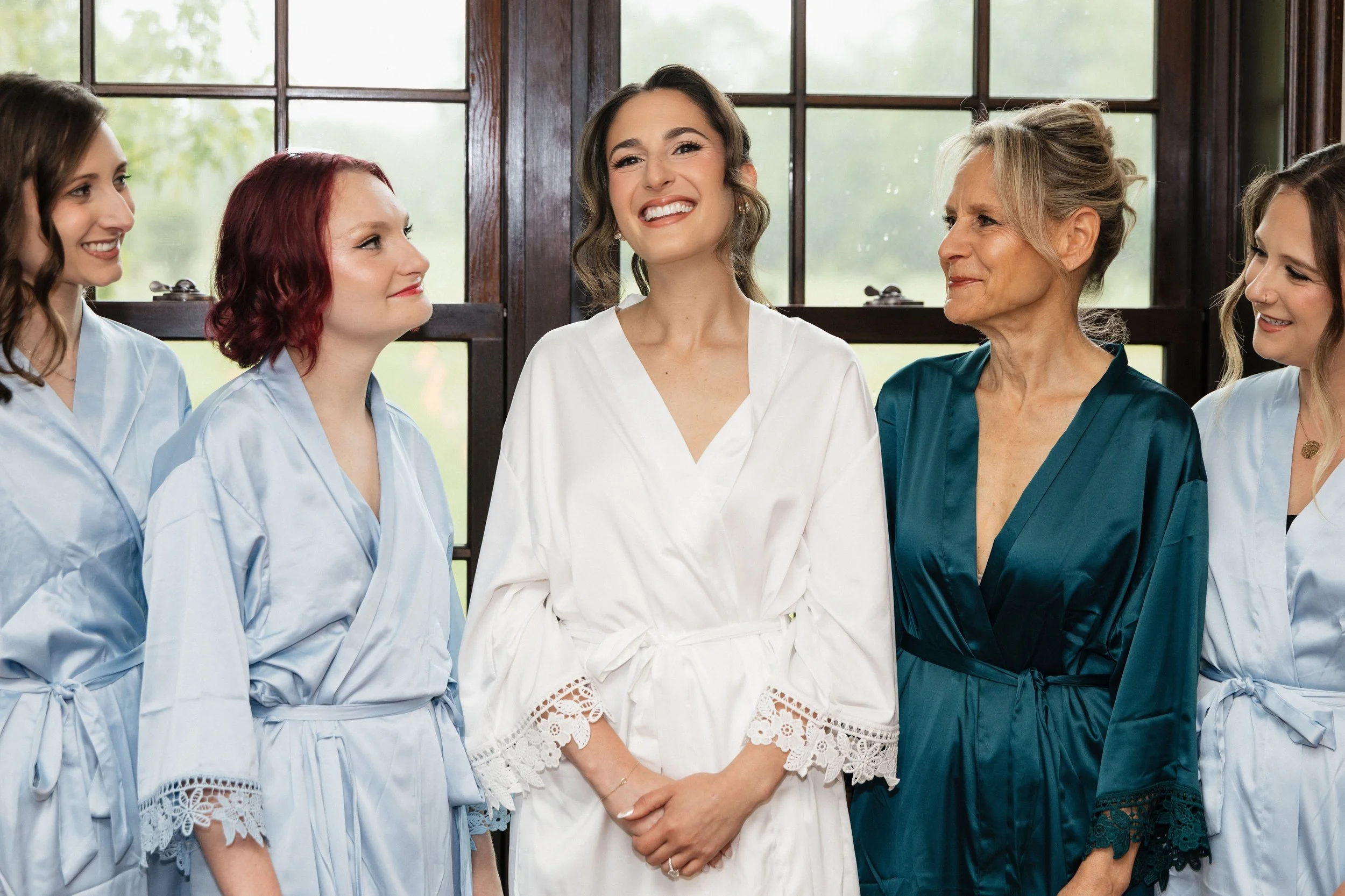 A group of women, some wearing light blue robes and one in a white robe, standing near a window with trees outside, smiling and engaging with each other in a bright, indoor setting.